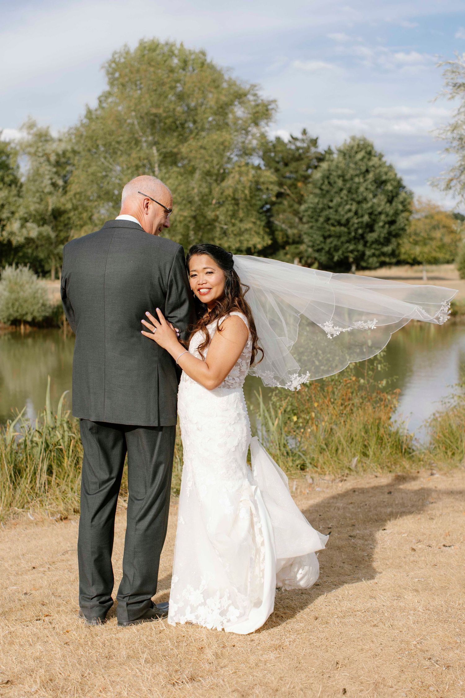 Bride looking over to the lake while the bride looks back at the camera, the vail is flowing in the breeze