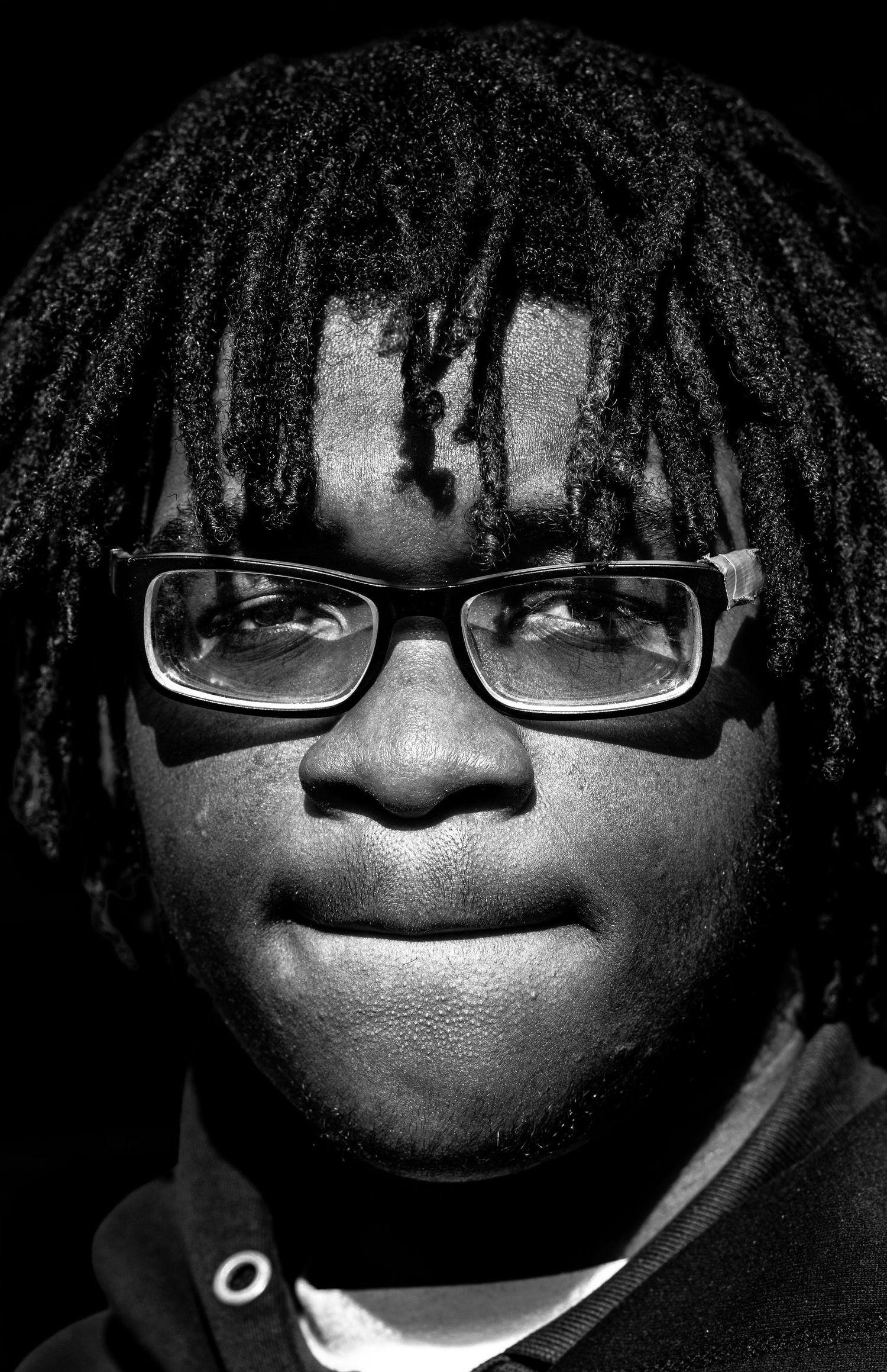 Black and white portrait of a young man with dreadlocks wearing rectangular glasses against dark background.