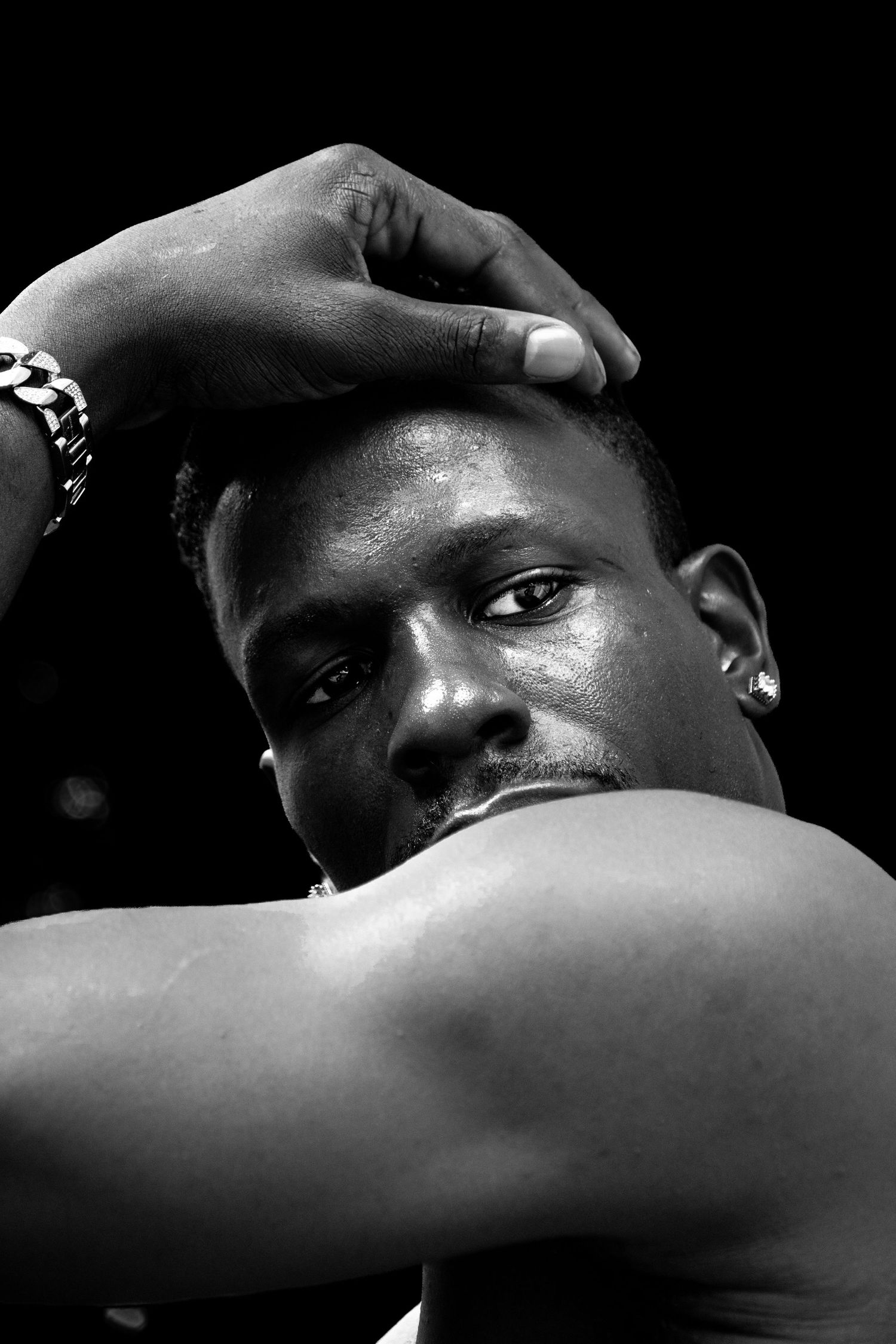 Black and white portrait of a muscular man with a bracelet resting his hand on his head against dark background.