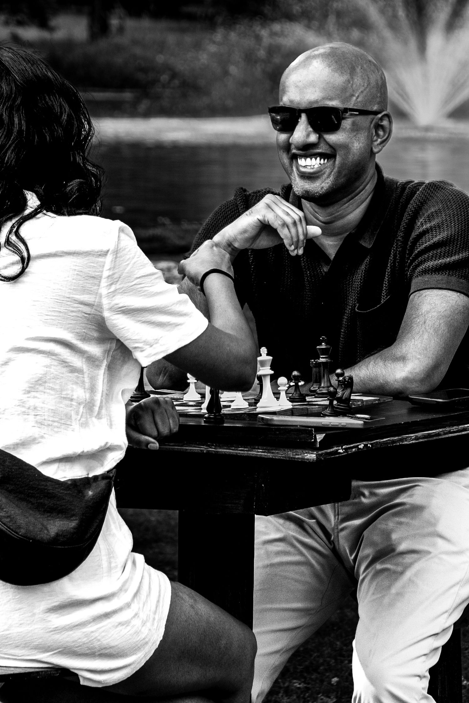 Black and white photo of a laughing bald man in sunglasses playing chess outdoors with a woman.