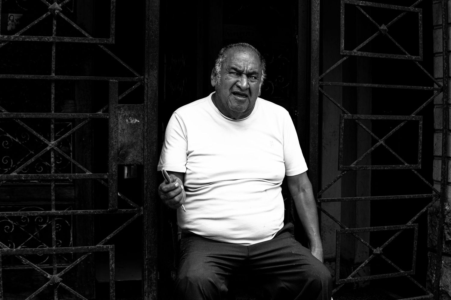 Black and white photo of a heavyset man in a white t-shirt sitting in a doorway.