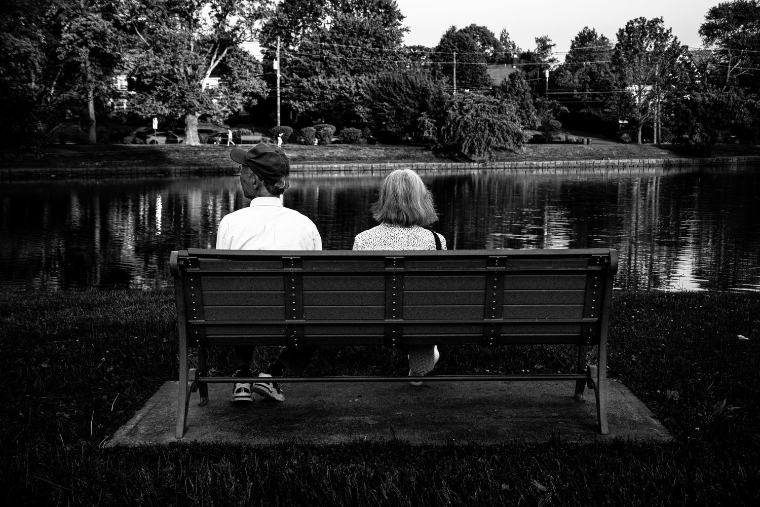 Black and white photo of a couple sitting on a park bench facing a calm reflective pond.
