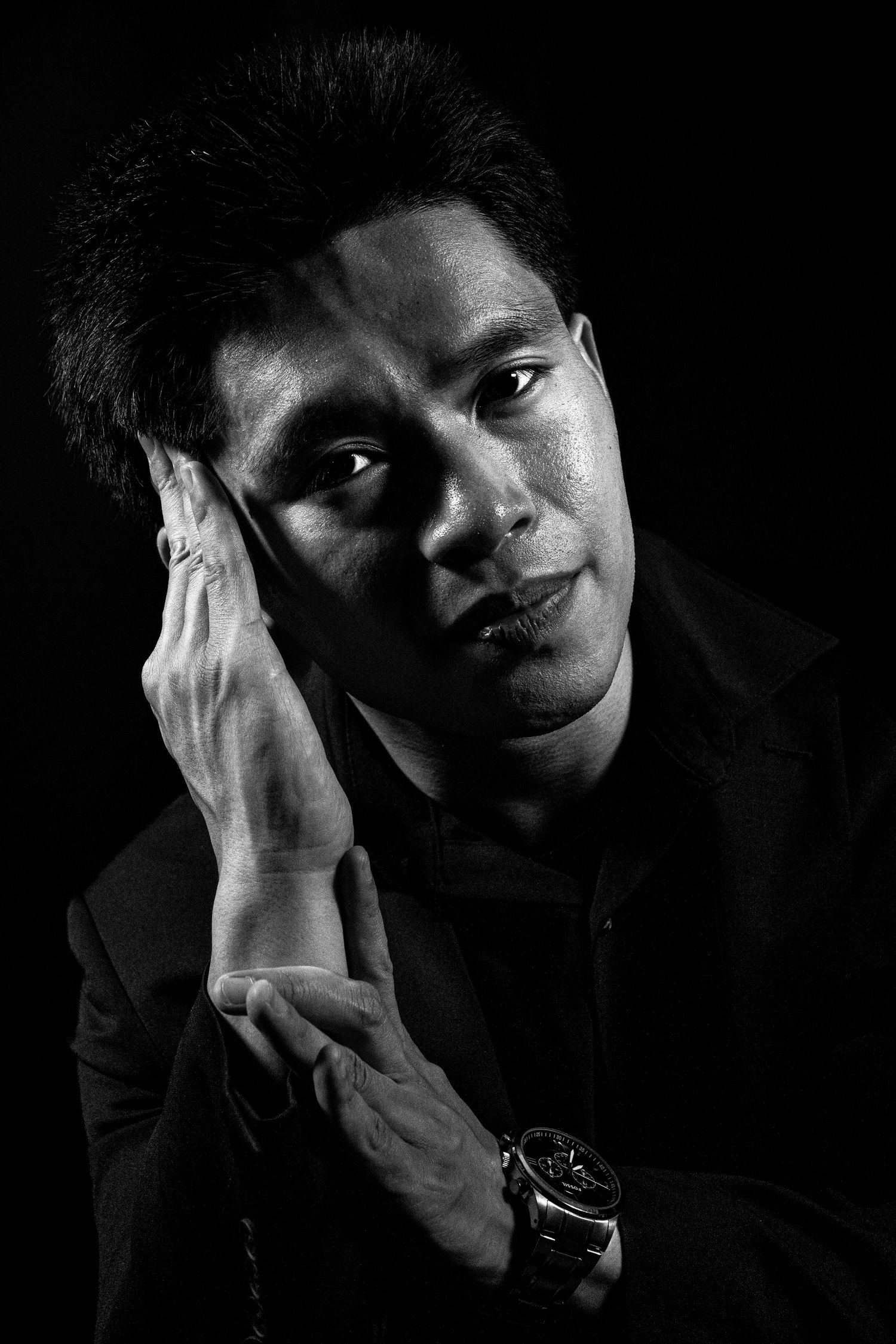 Black and white portrait of a thoughtful man in a dark shirt resting his hand on his cheek, wearing a watch.