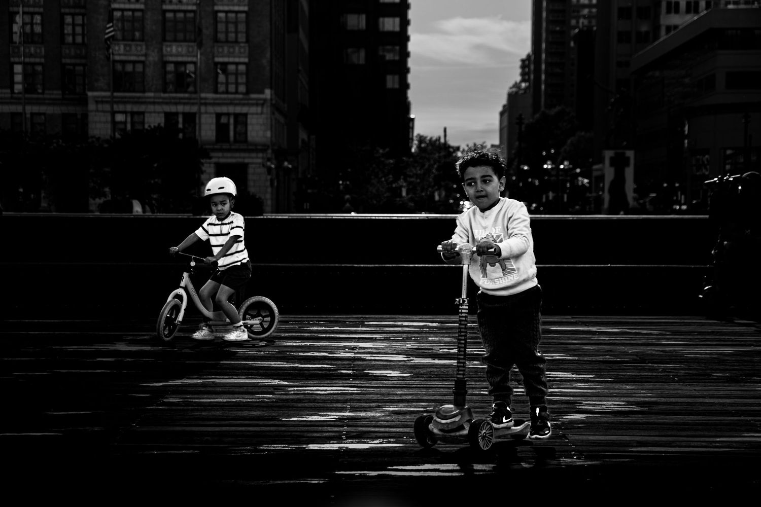 Two children riding scooters and balance bikes on a city street in black and white photography.