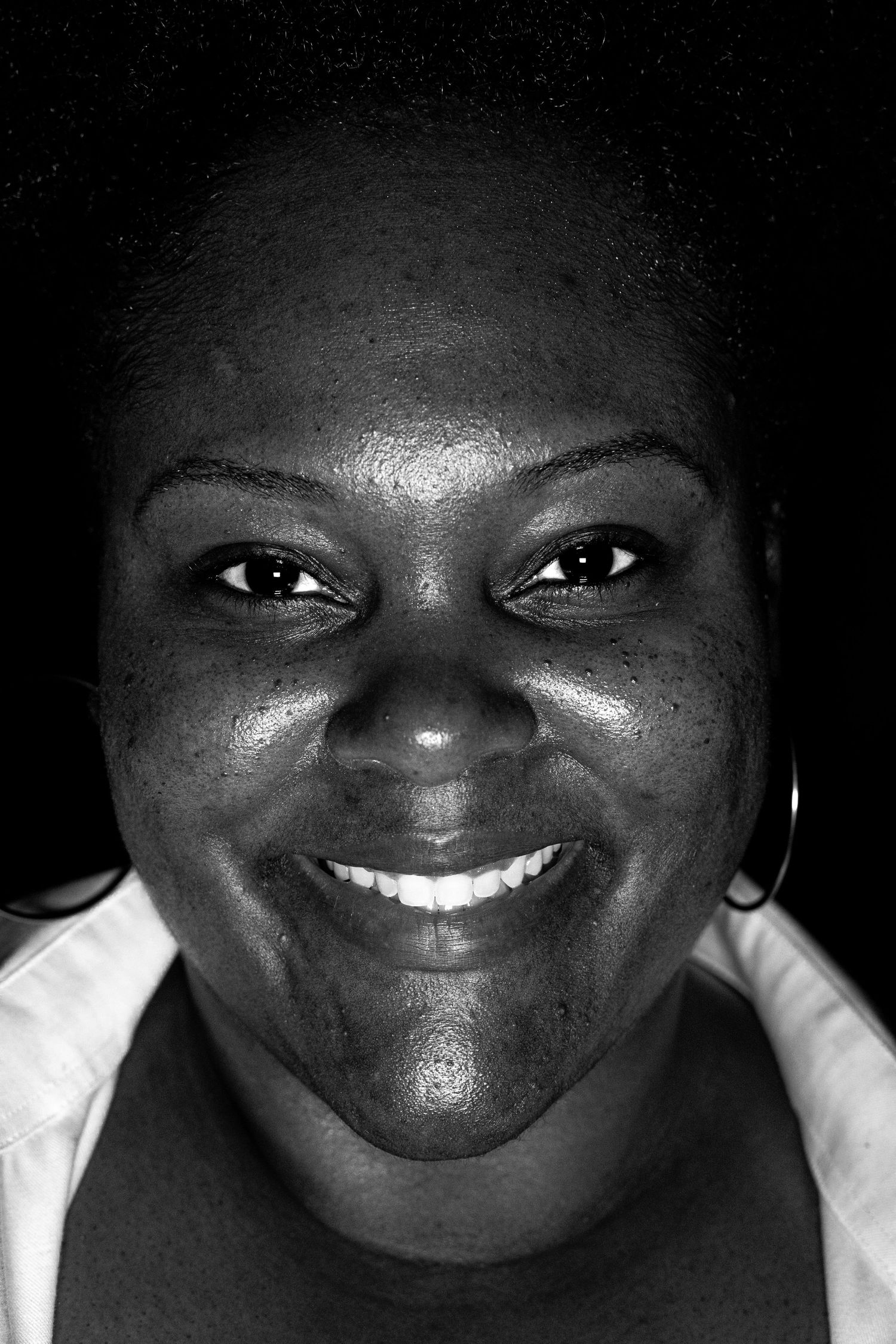 Black and white close-up portrait of a smiling woman with hoop earrings against dark background.