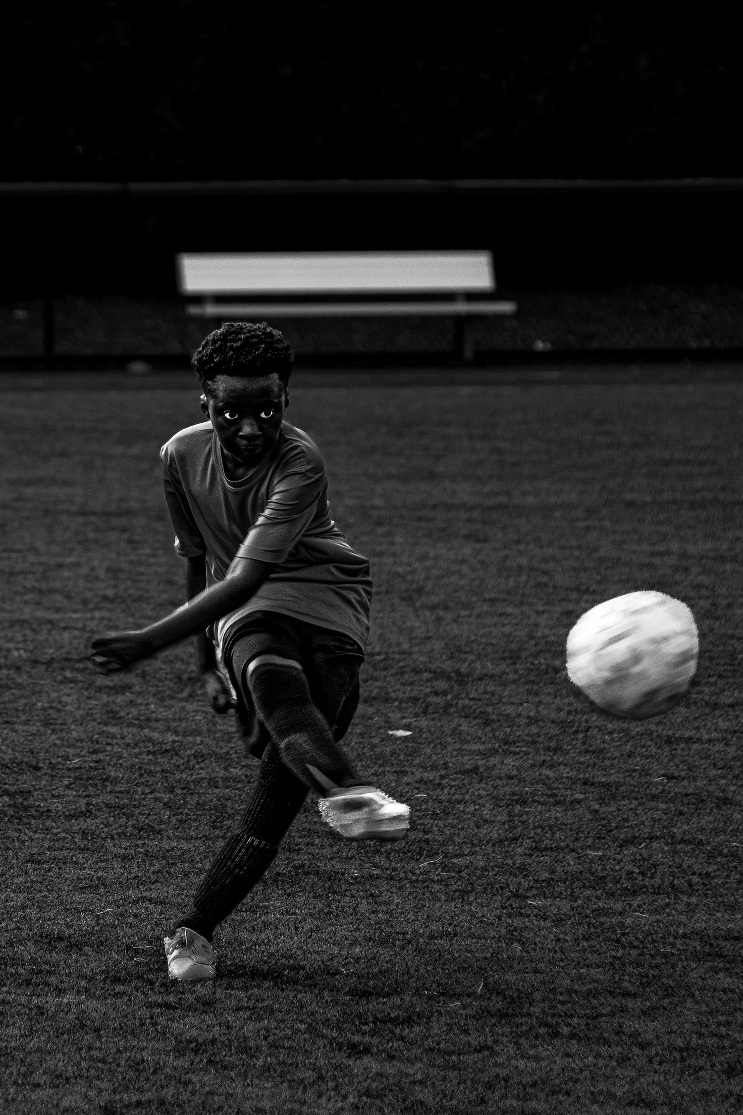 Black and white photo of young soccer player kicking ball on field at night.