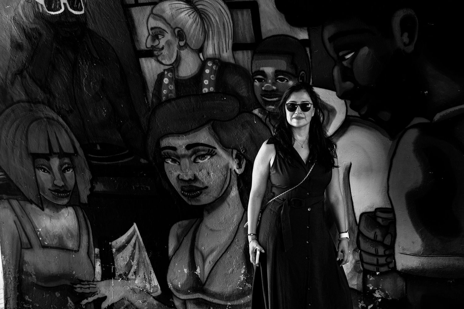 Black and white photo of a woman in sunglasses standing in front of a colorful mural.