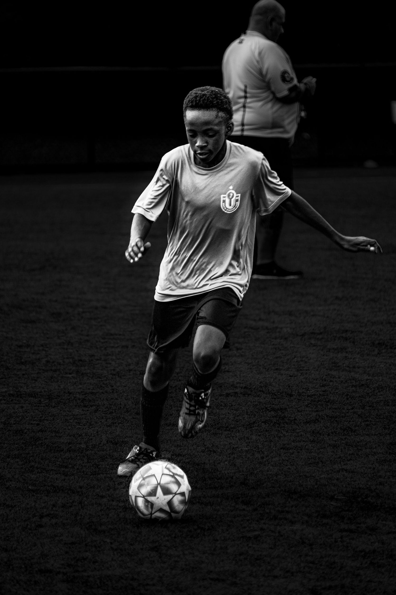 Young black and white photo of boy dribbling soccer ball on field during practice.