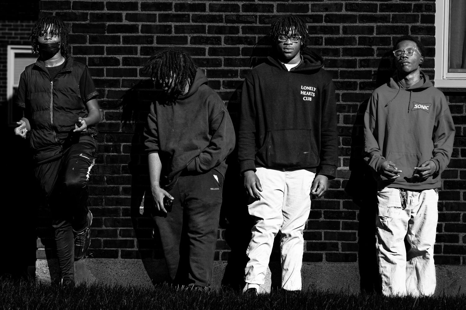 Black and white photo of four young men leaning against a brick wall outdoors.