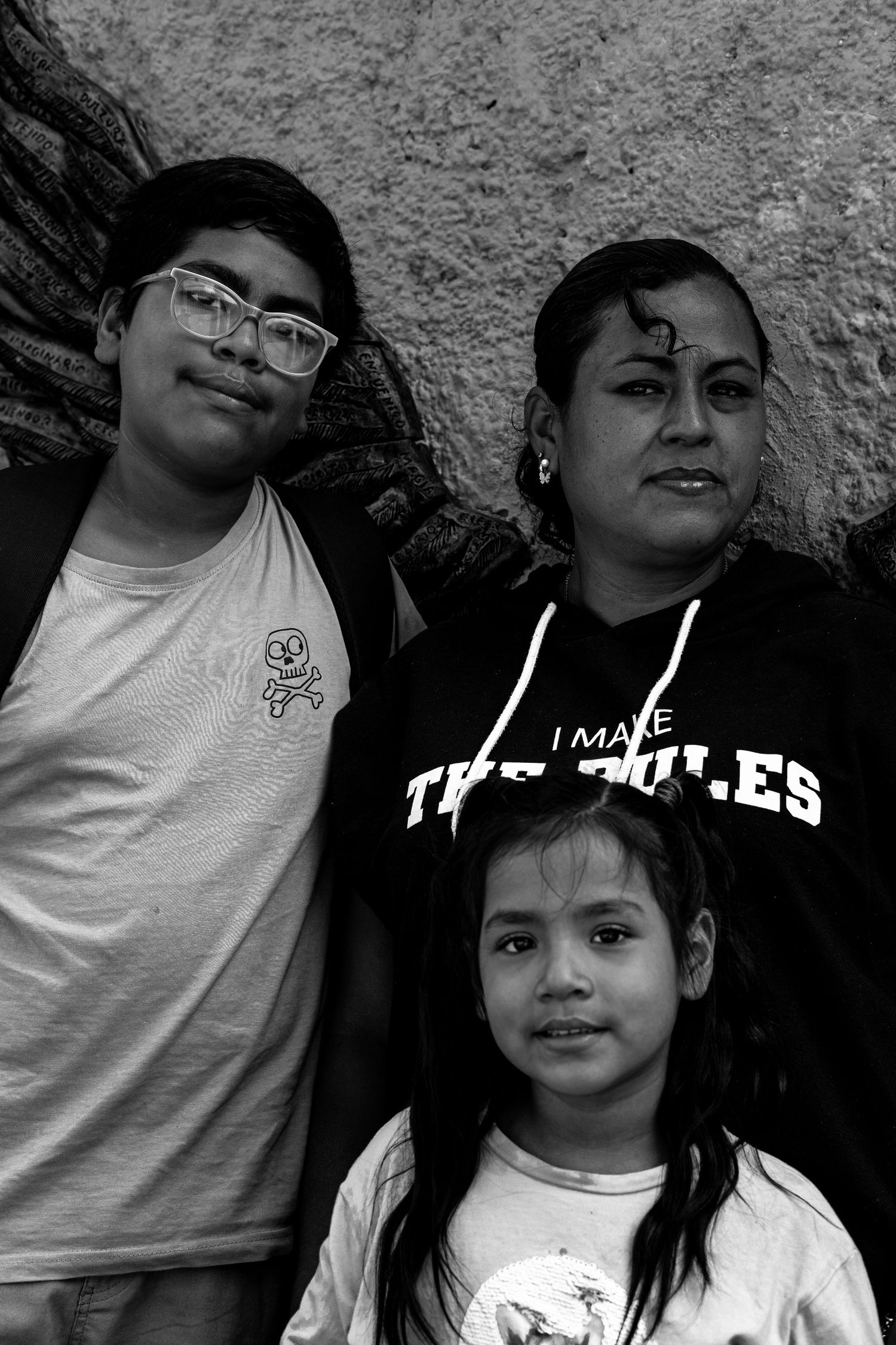 Black and white portrait of a woman with two children standing against a textured wall.