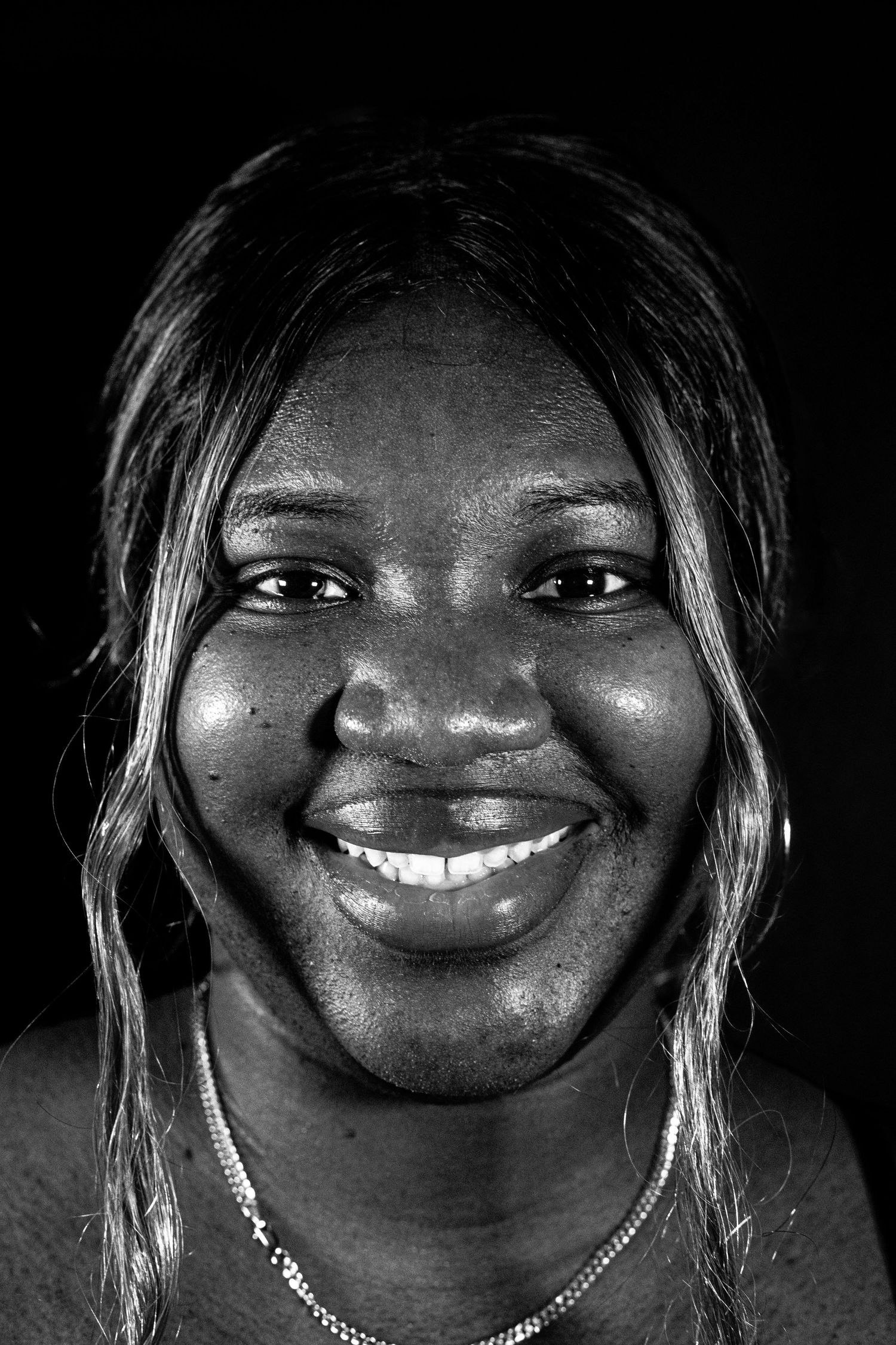 Black and white portrait of a smiling woman with long hair wearing a necklace against dark background.