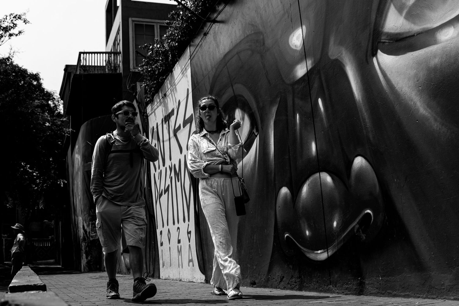 Black and white photo of two people walking past a large graffiti mural on a city street.