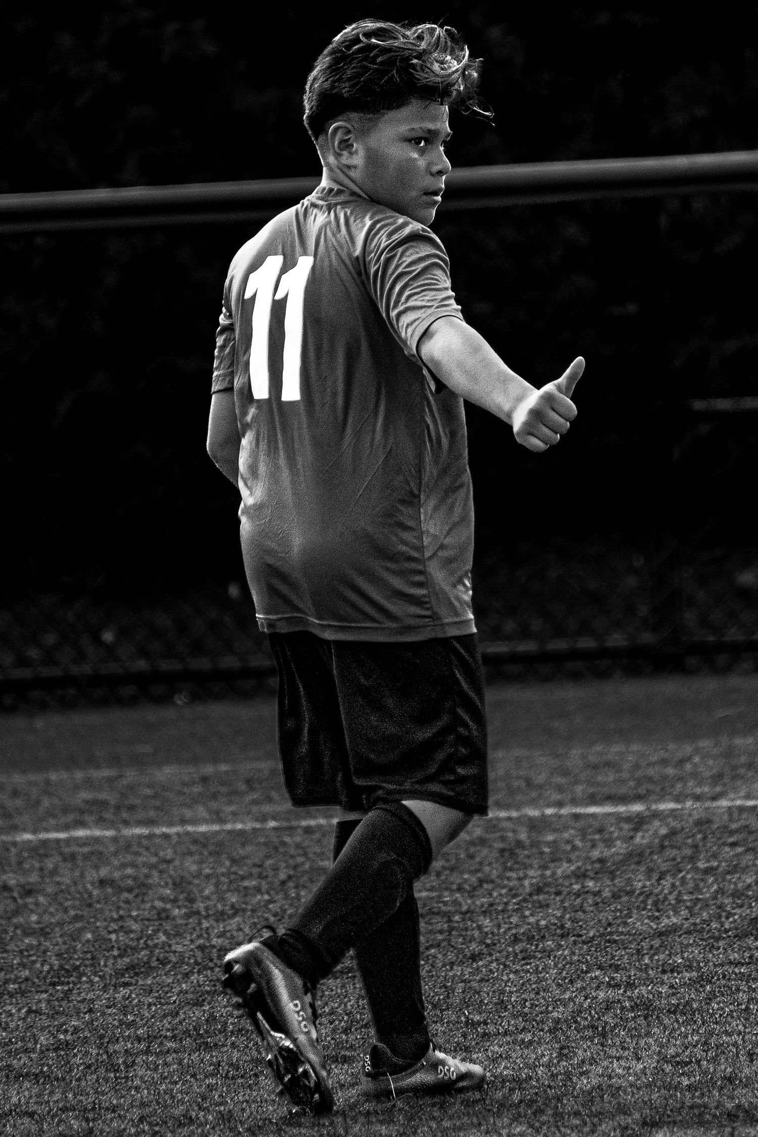 Young soccer player wearing number 11 jersey giving thumbs up on field in black and white.