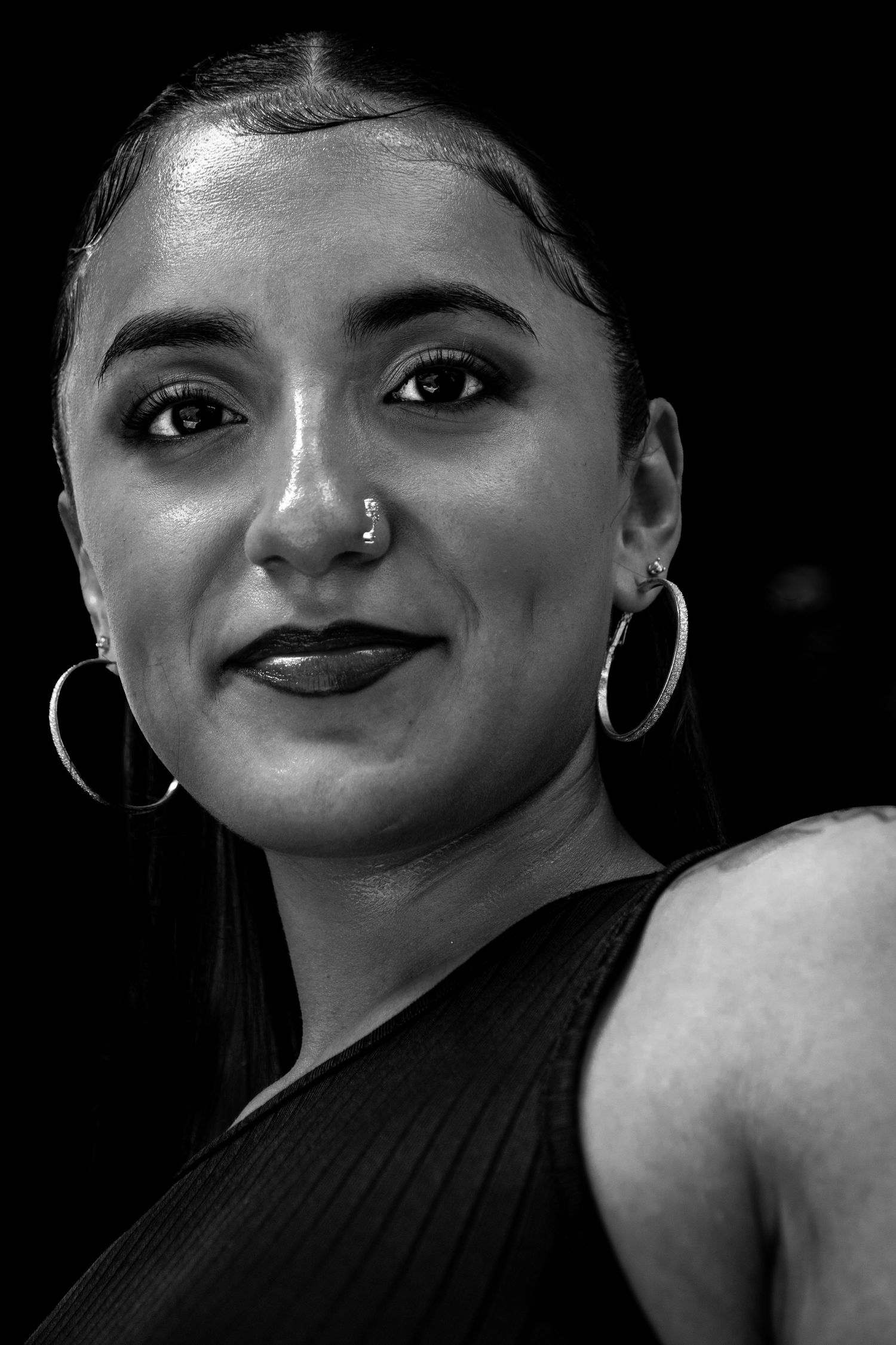Black and white portrait of a young woman with hoop earrings and nose ring, smiling softly against dark background.