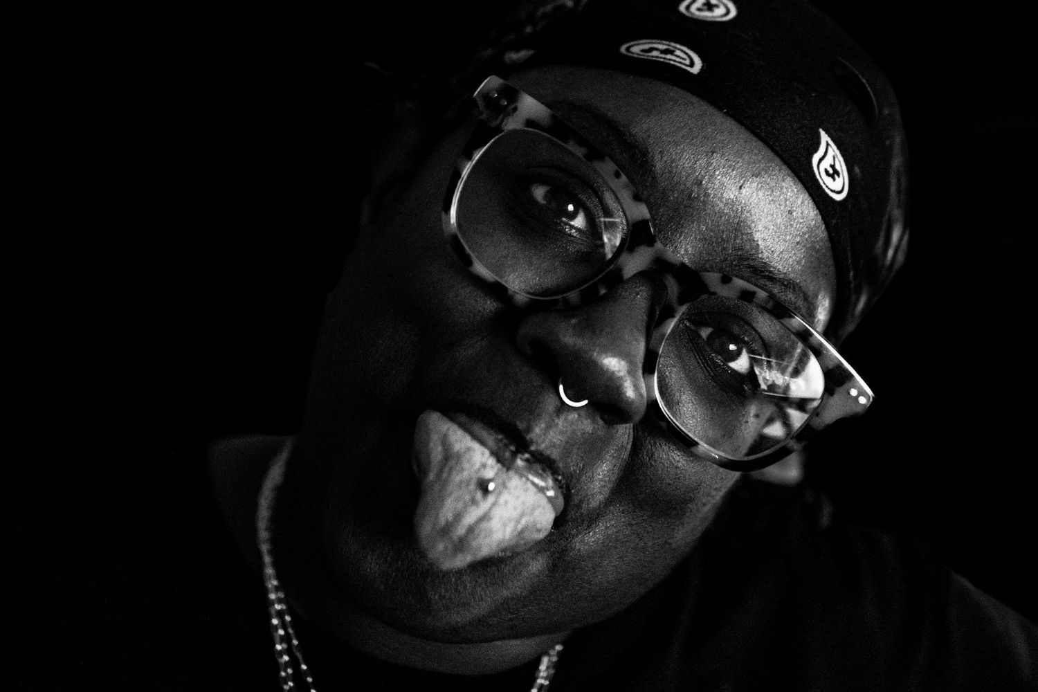 Black and white close-up portrait of a person wearing glasses and a cap sticking out tongue.