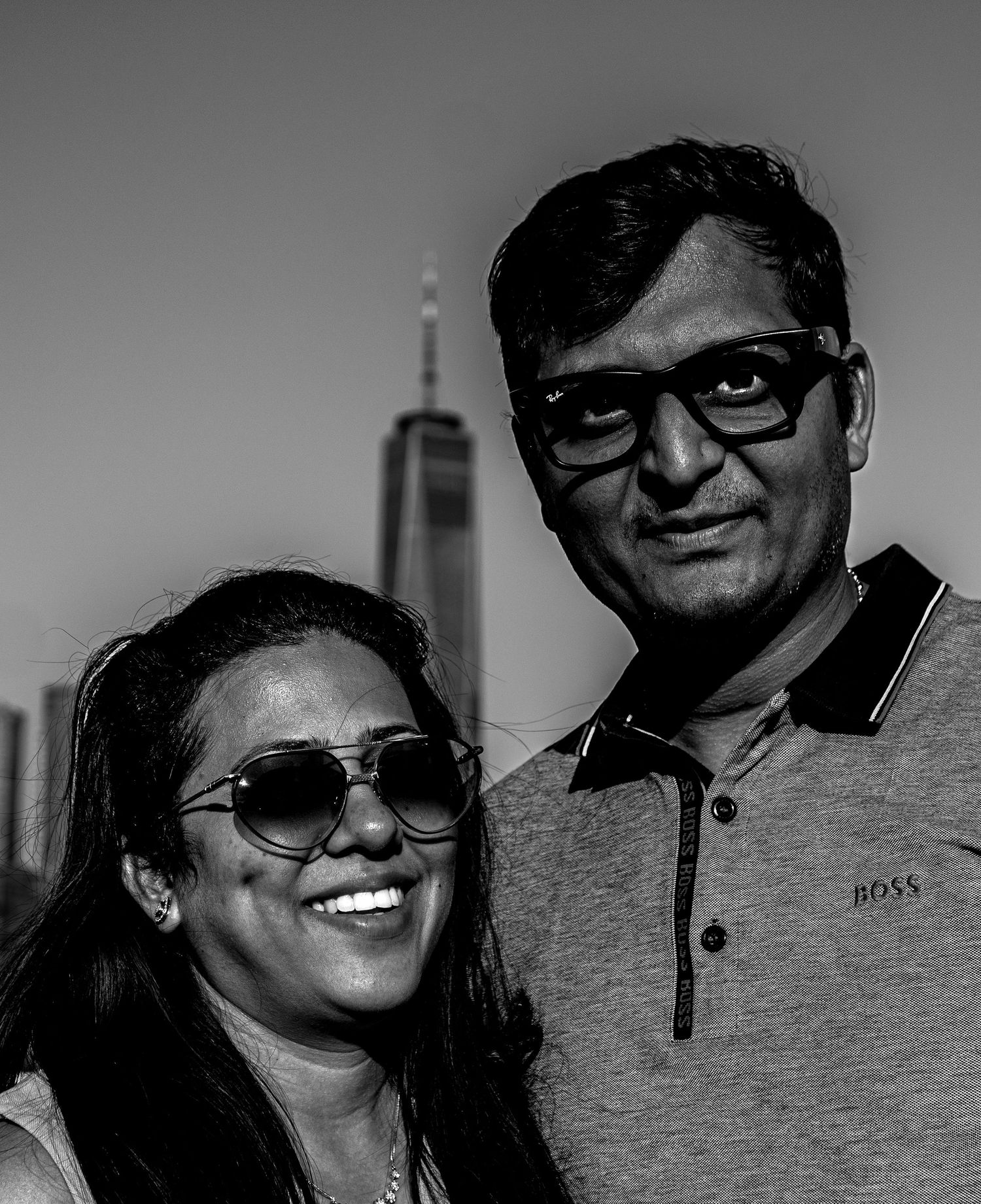 Black and white portrait of a smiling couple wearing sunglasses with a city skyline behind them.