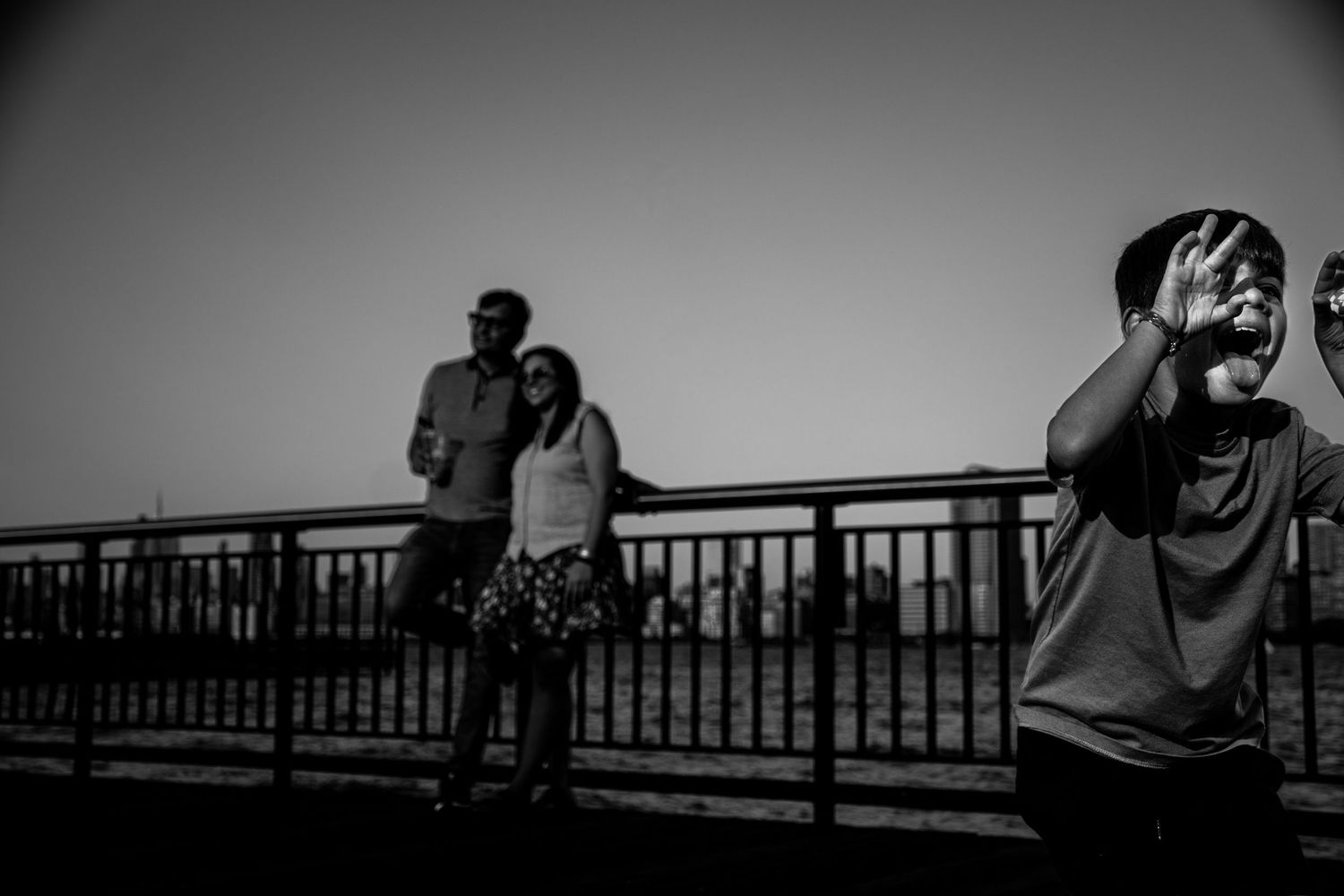 Child poses for camera while couple stands on bridge in black and white photo.