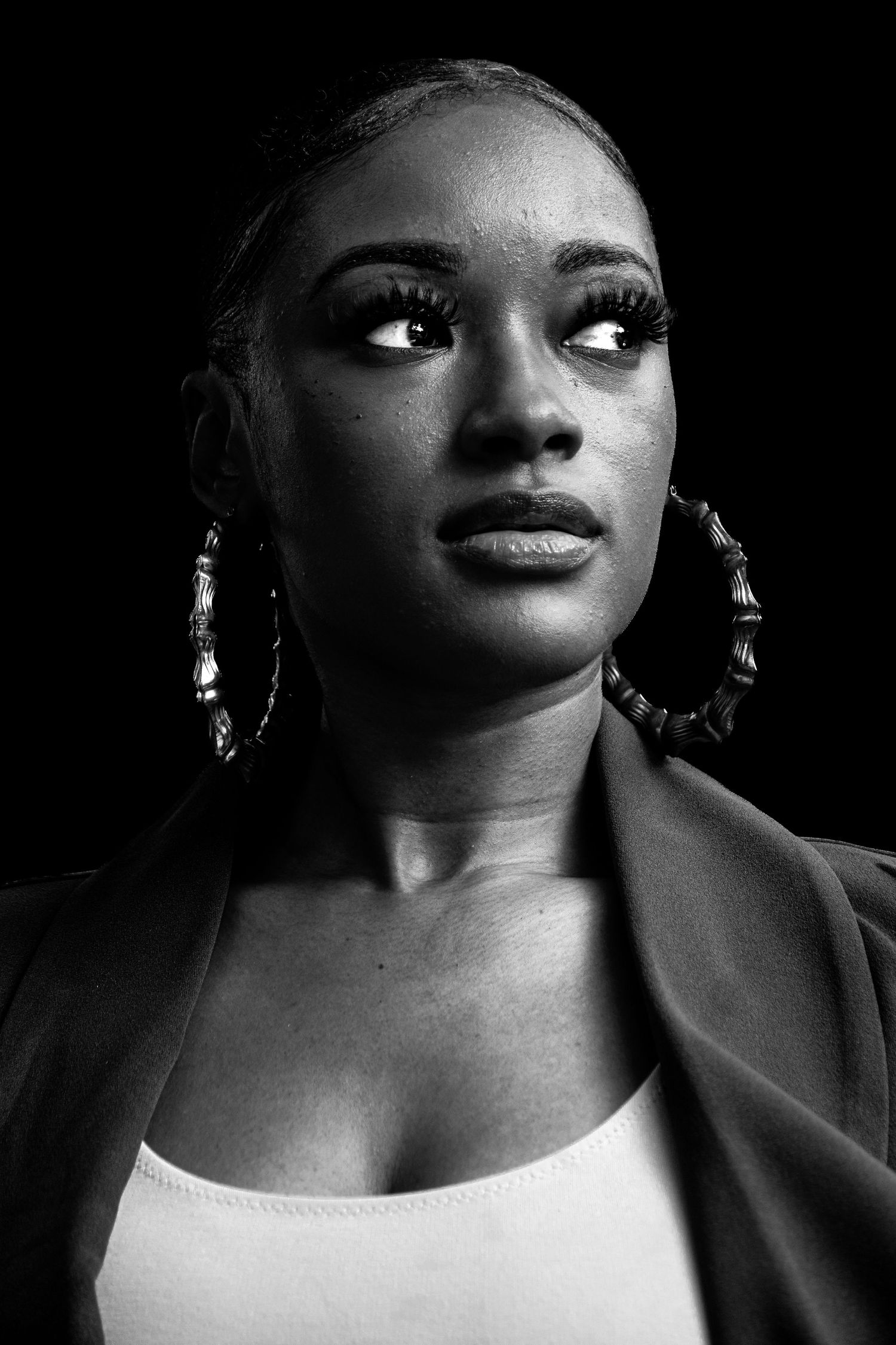 Black and white portrait of a young woman wearing hoop earrings and a blazer, gazing upward against dark background.