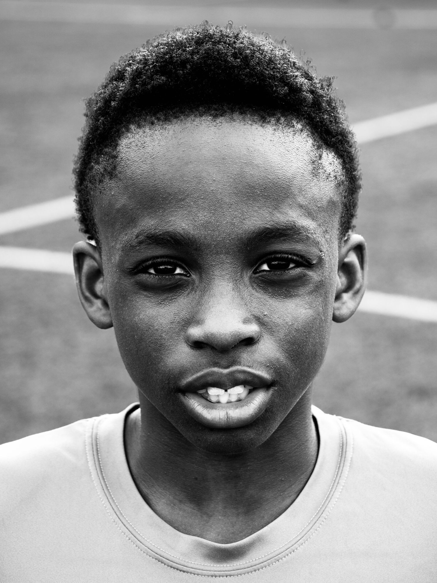 Black and white close-up portrait of a young boy with short curly hair on a sports field.