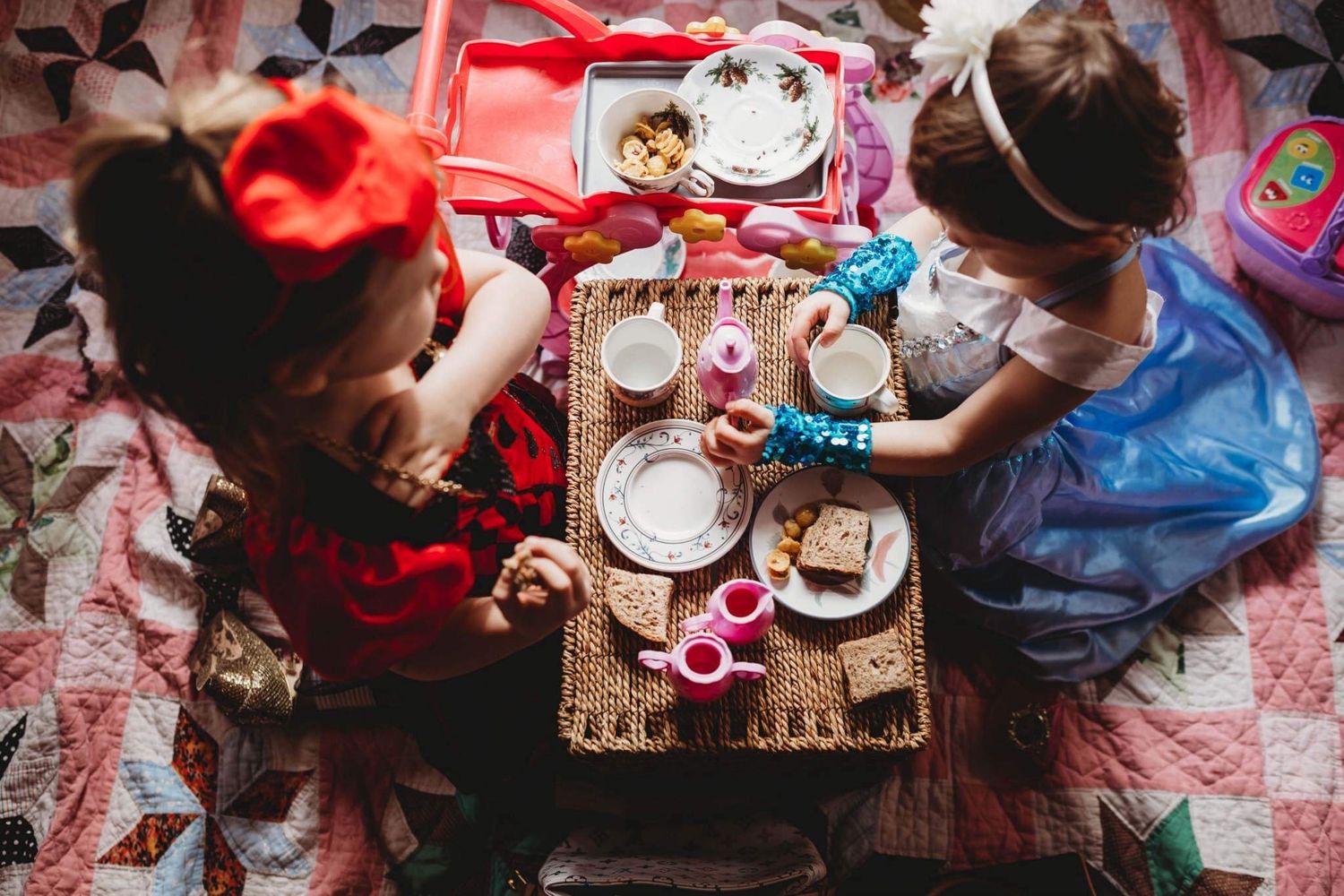 Two girls in princess costumes having a tea party on a quilt from above.