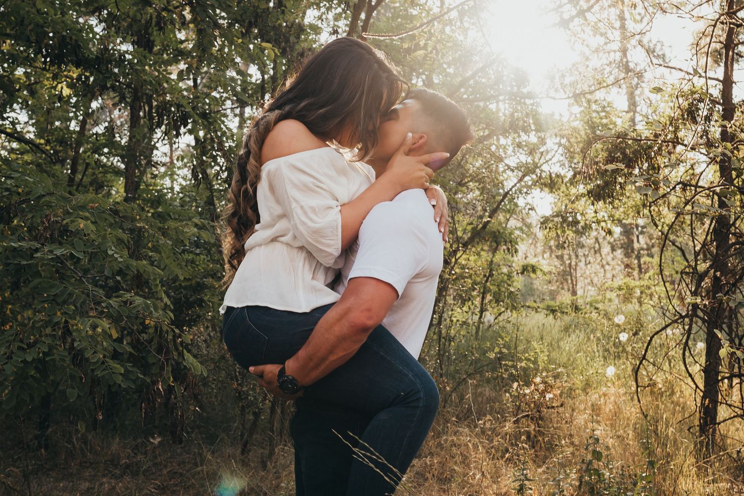 Romantic couple kissing in a sunlit forest, man lifting woman in white blouse among green trees.