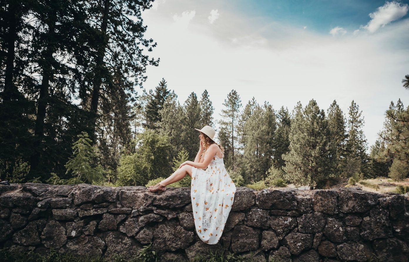Woman in white floral dress and hat sitting on stone wall surrounded by tall pine trees.