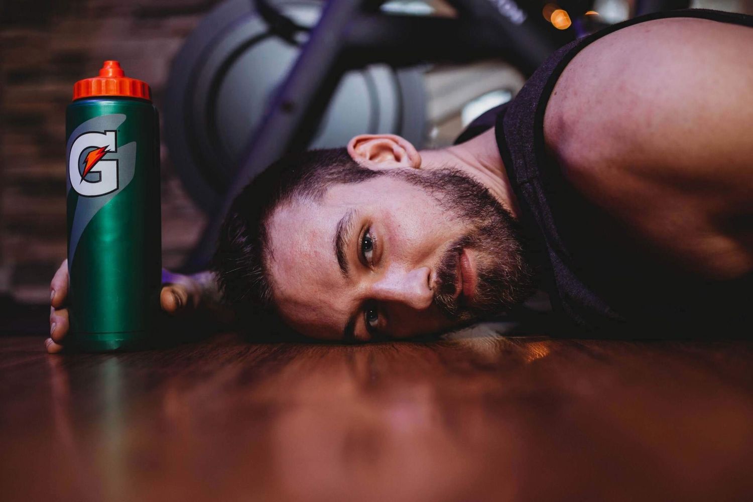 Exhausted bearded man lying on wooden floor next to a green Gatorade bottle after workout.