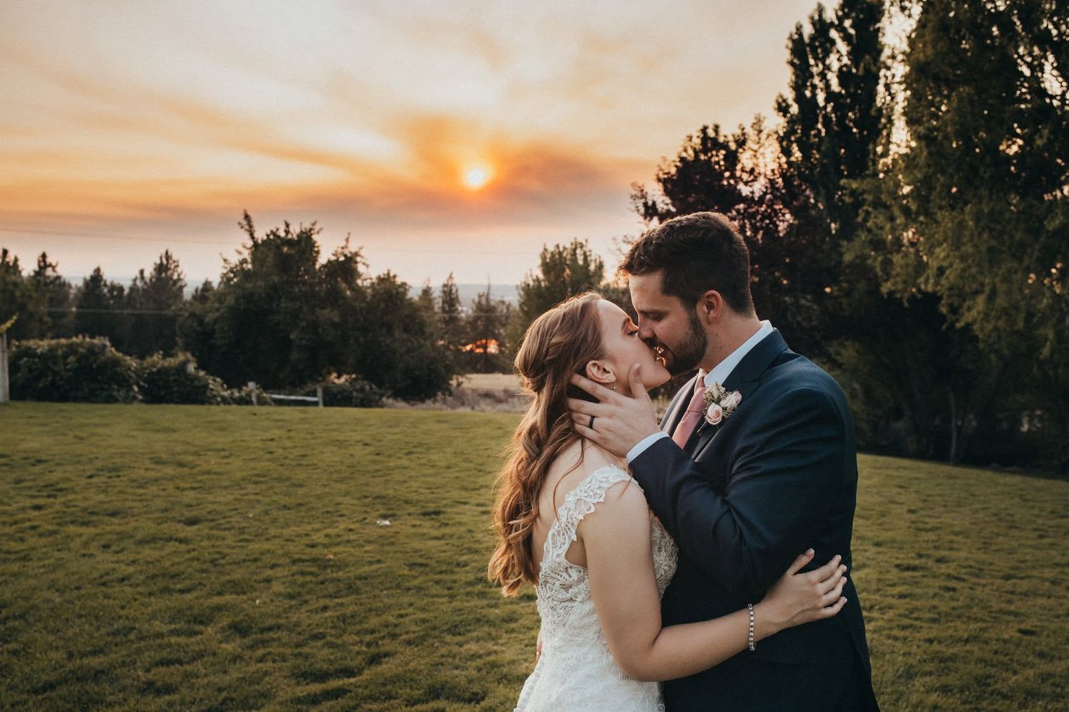 Bride and groom share a romantic kiss at sunset on a lush green lawn at wedding.