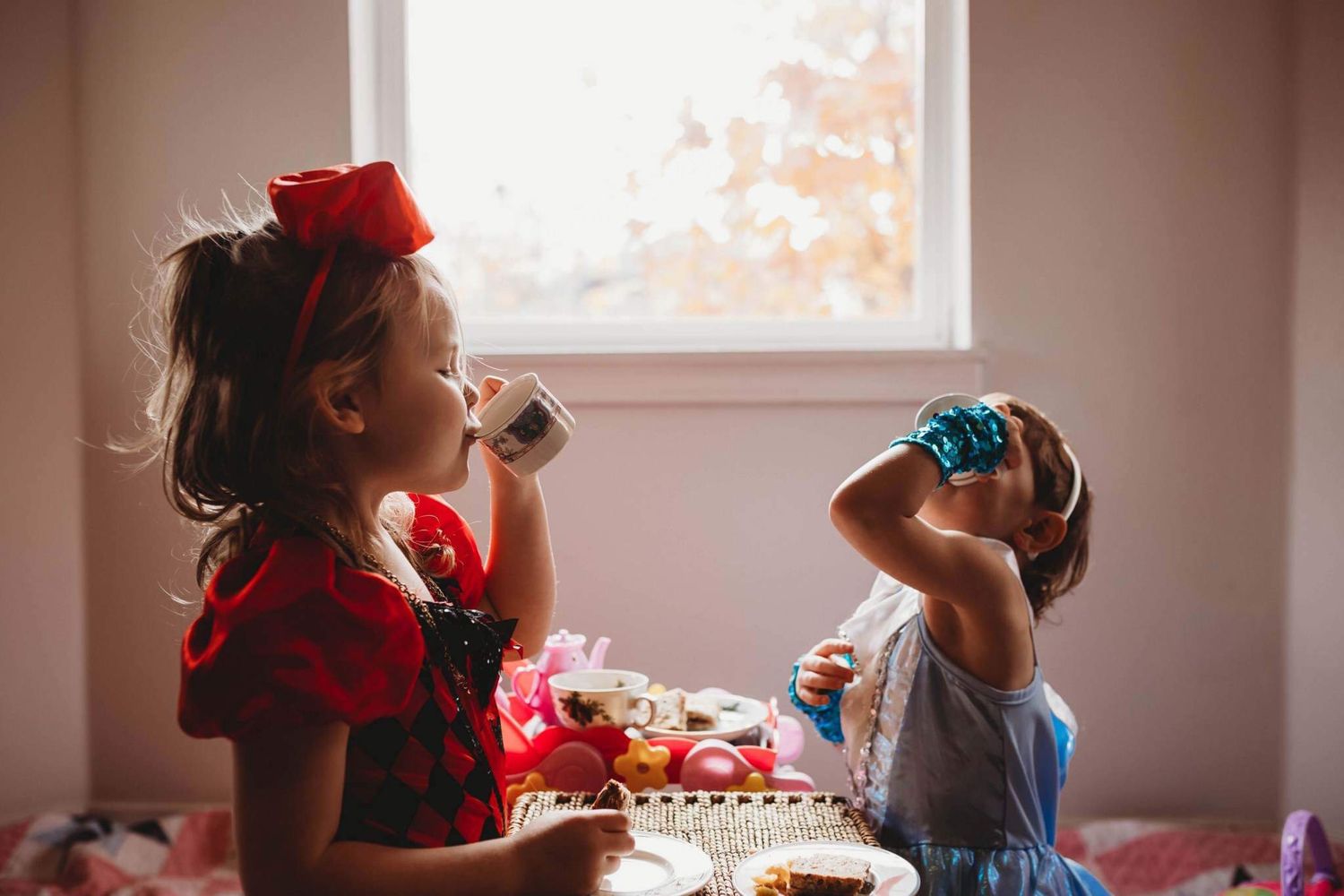 Two young girls in costumes having a pretend tea party indoors near a bright window.