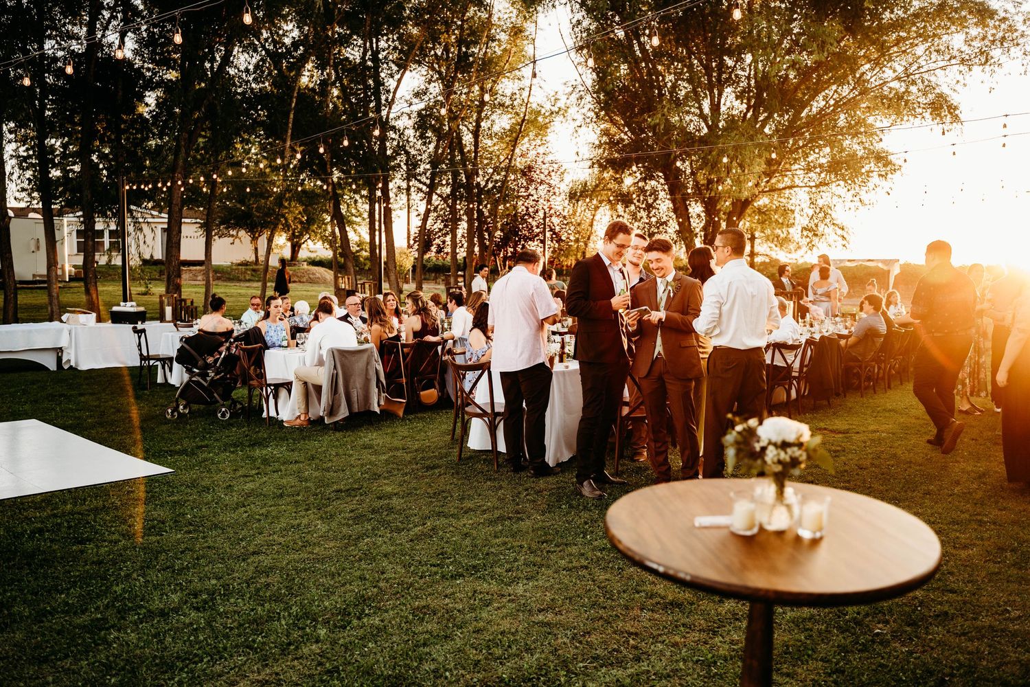 Wedding reception guests mingle on a golden-lit lawn surrounded by string lights at dusk.
