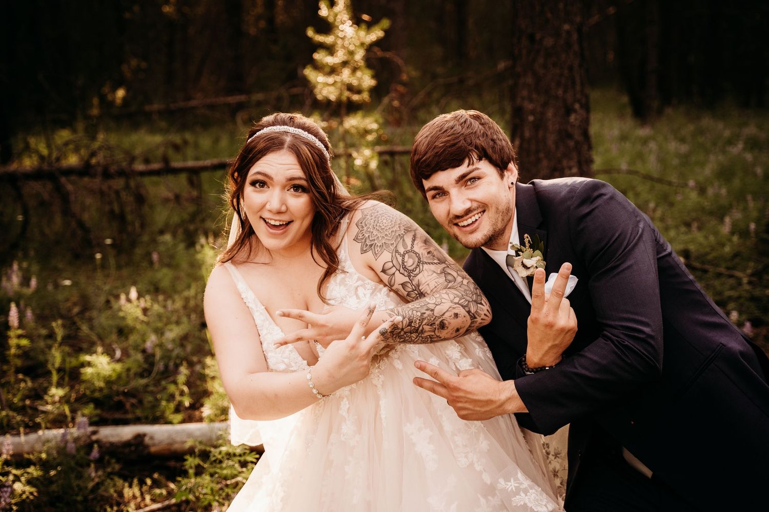 Laughing bride in lace gown and groom make playful gestures during fun outdoor forest wedding portraits.