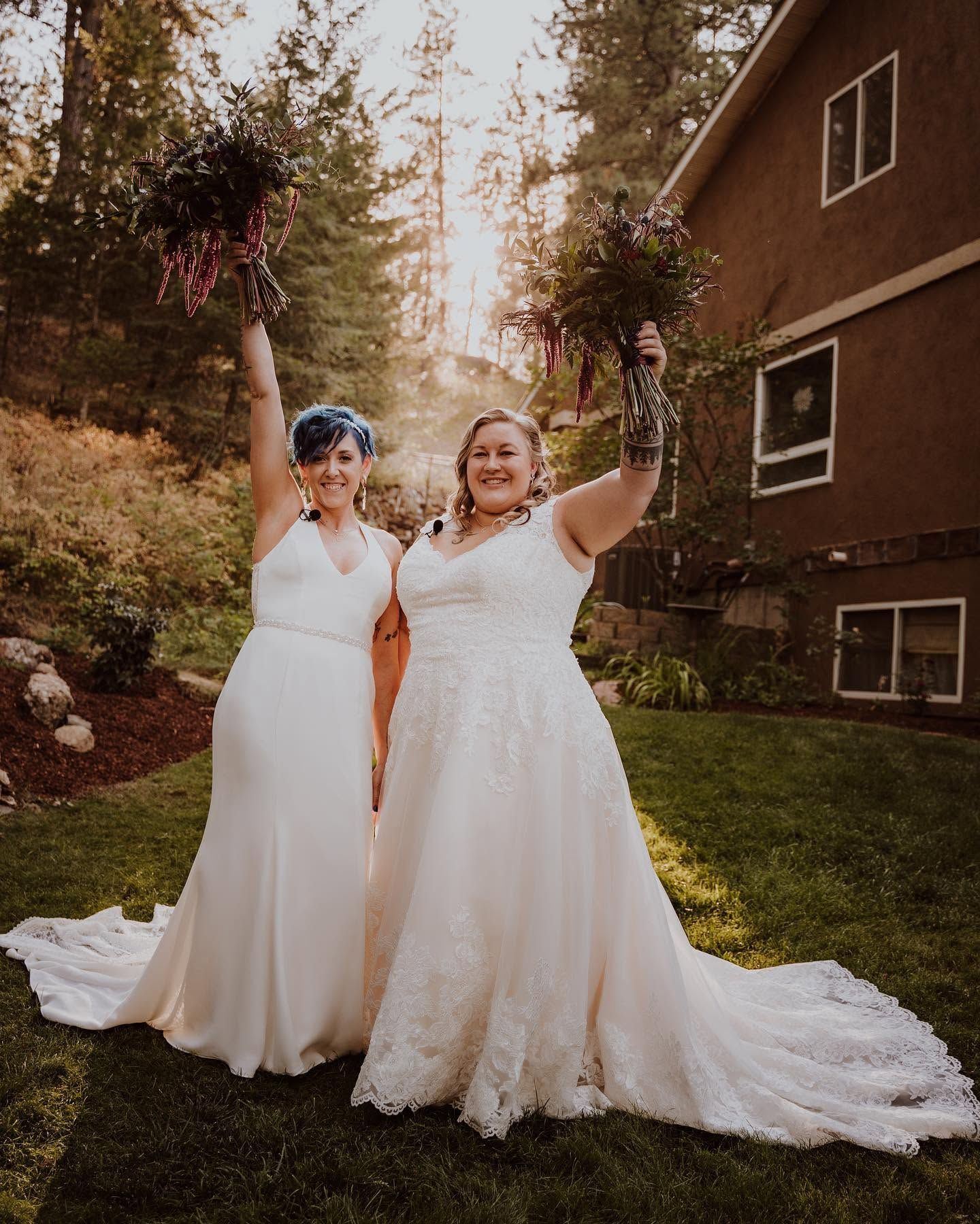 Two brides in white wedding gowns joyfully raise their bouquets in the air at golden hour outdoors.