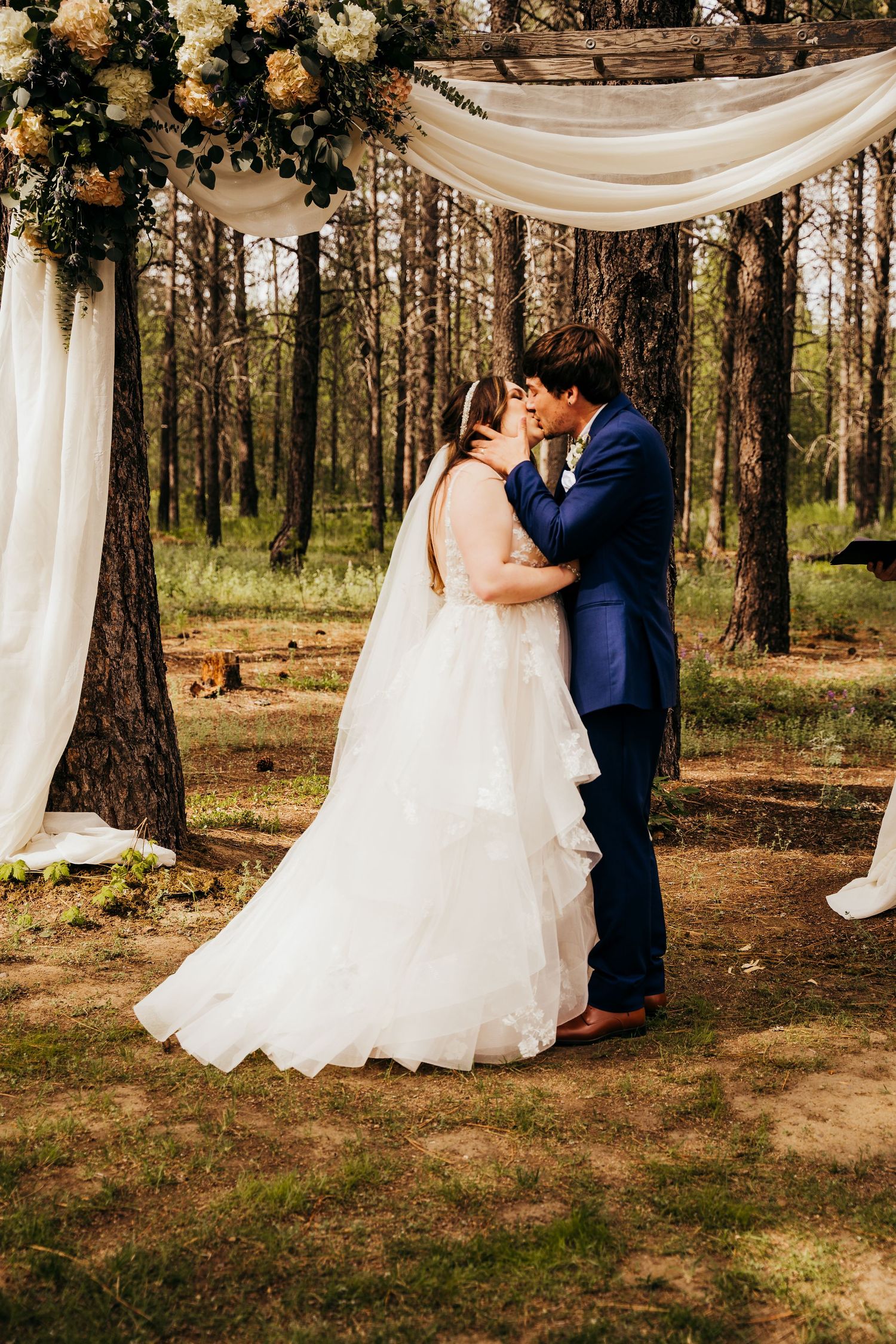 Bride and groom share their first kiss under a draped floral arch in a pine forest.