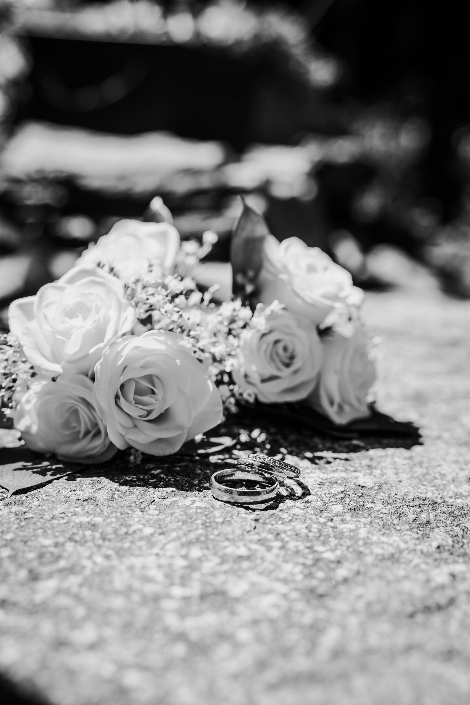 Wedding rings rest beside white rose bouquet on stone in black and white photo.