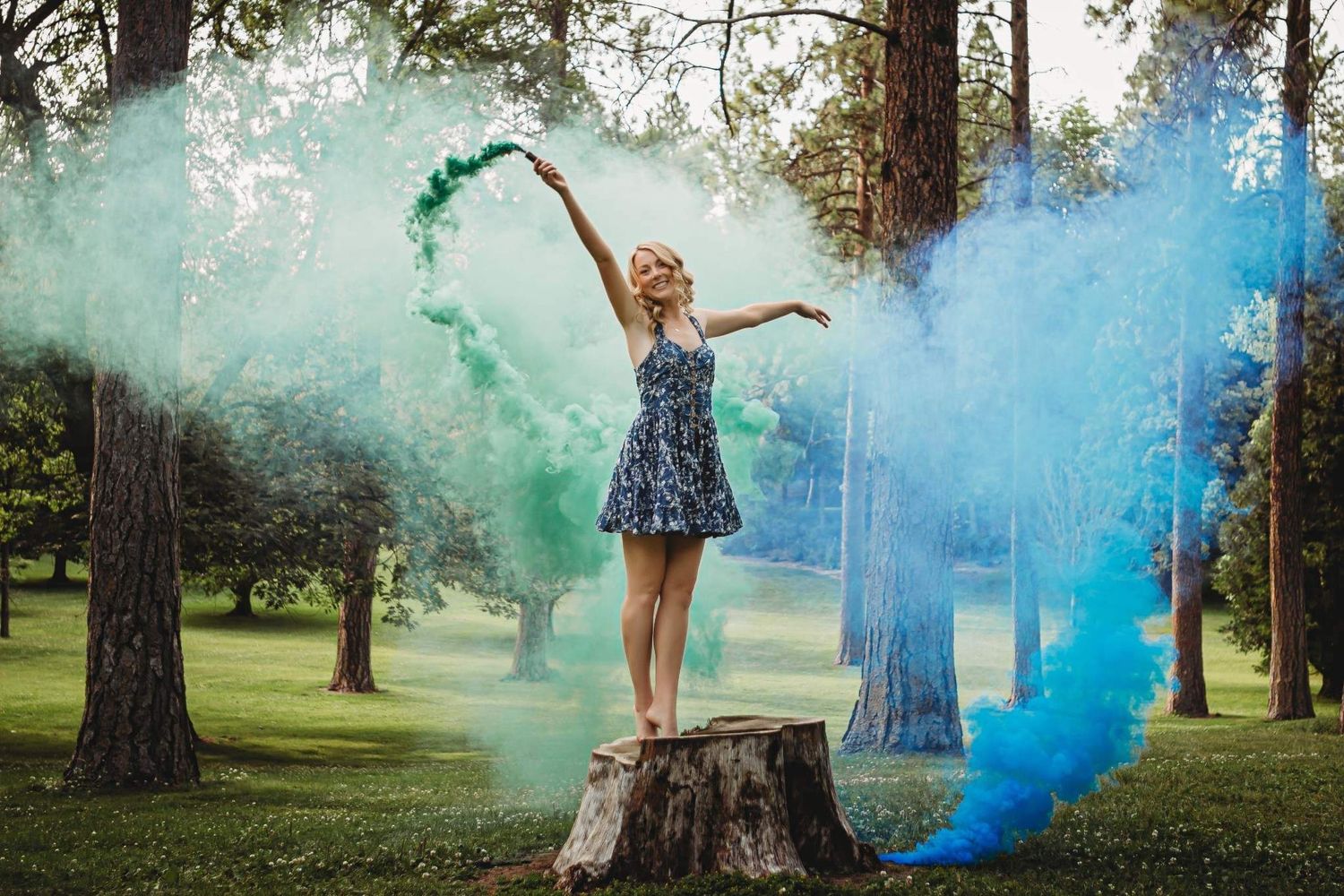 Blonde woman in floral dress standing on tree stump with colorful smoke bombs in park.