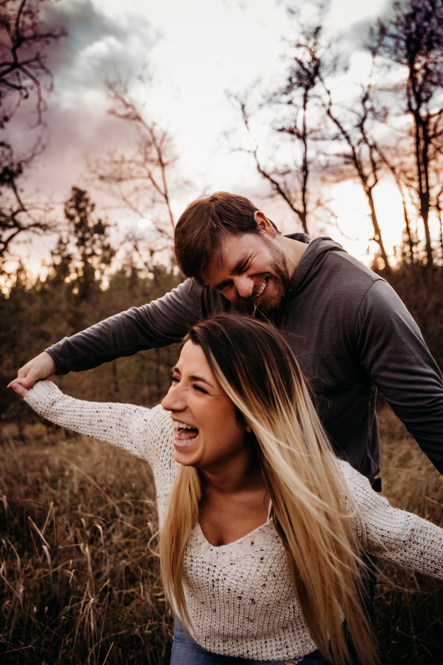 Couple laughing and playing in a field at sunset with trees in the background.