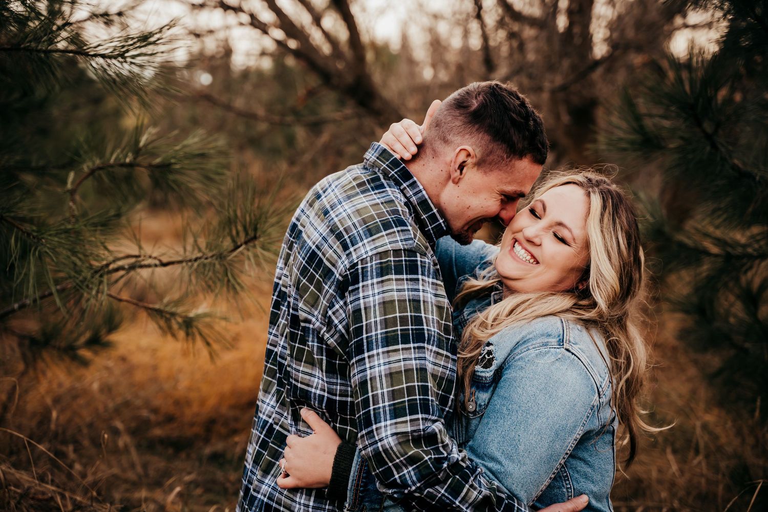 Couple hugging warmly among pine trees in a cozy flannel outdoor fall portrait.
