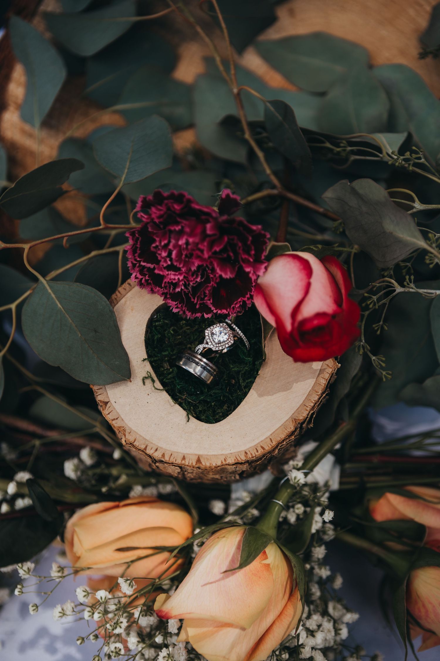 Wedding rings displayed on a wooden slice surrounded by roses and eucalyptus greenery.