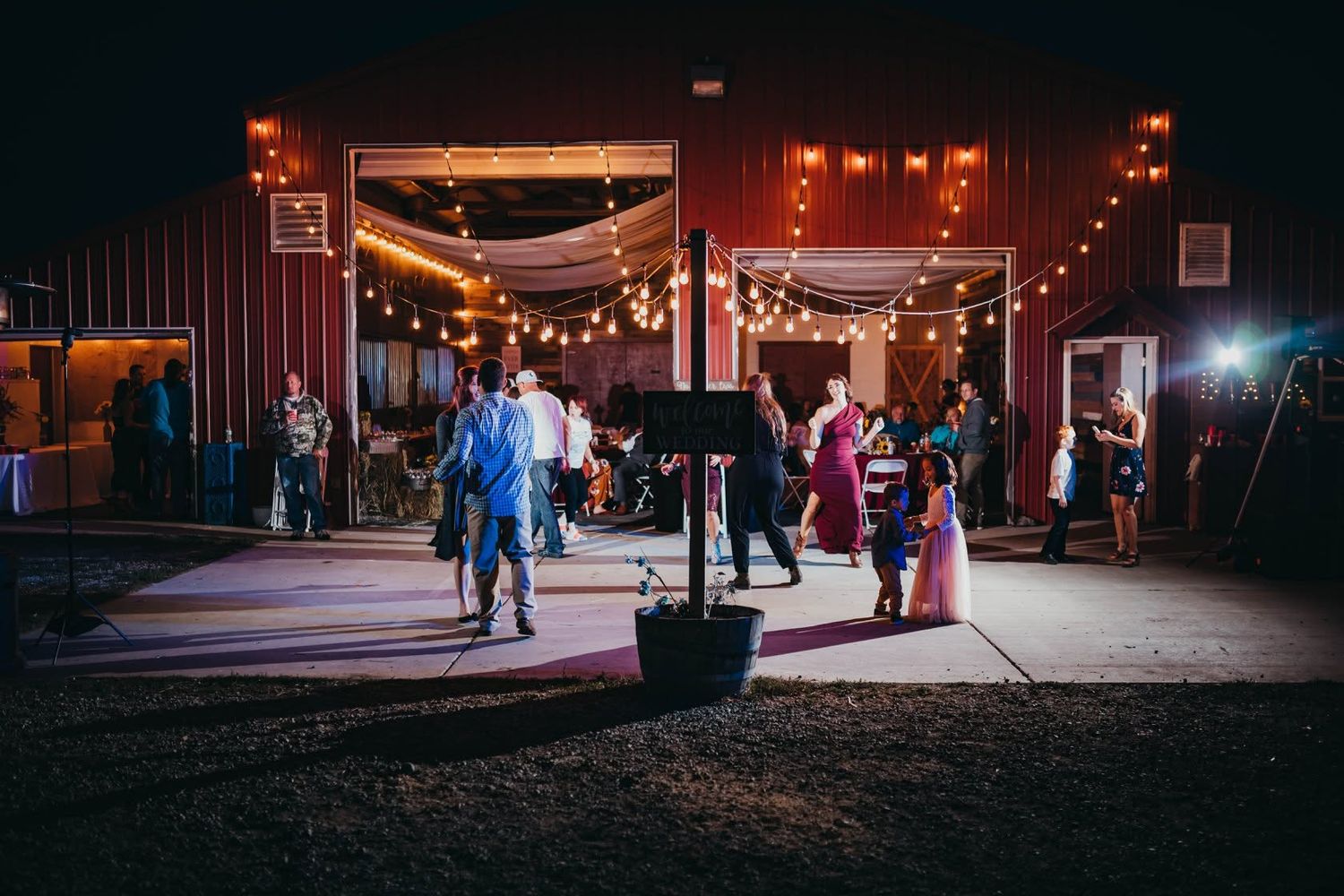 Wedding guests mingle and dance outside a rustic red barn venue under string lights at night.