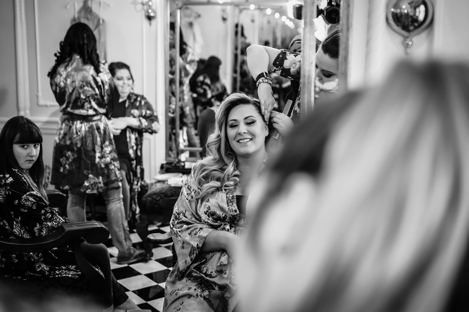 Bride smiles during hair and makeup preparation with bridesmaids in floral robes.