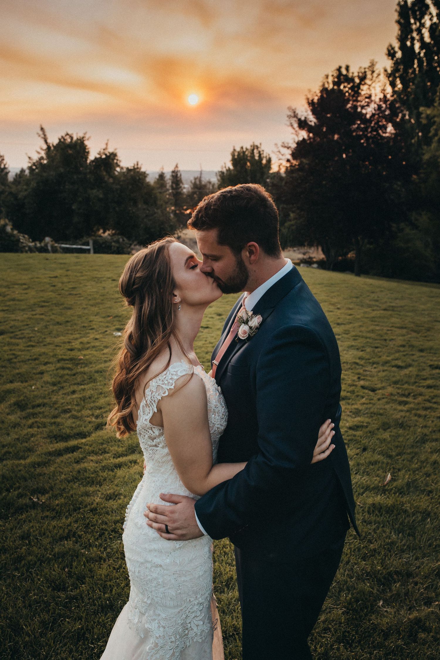 Newlyweds share a passionate kiss against a stunning sunset sky on their wedding day.
