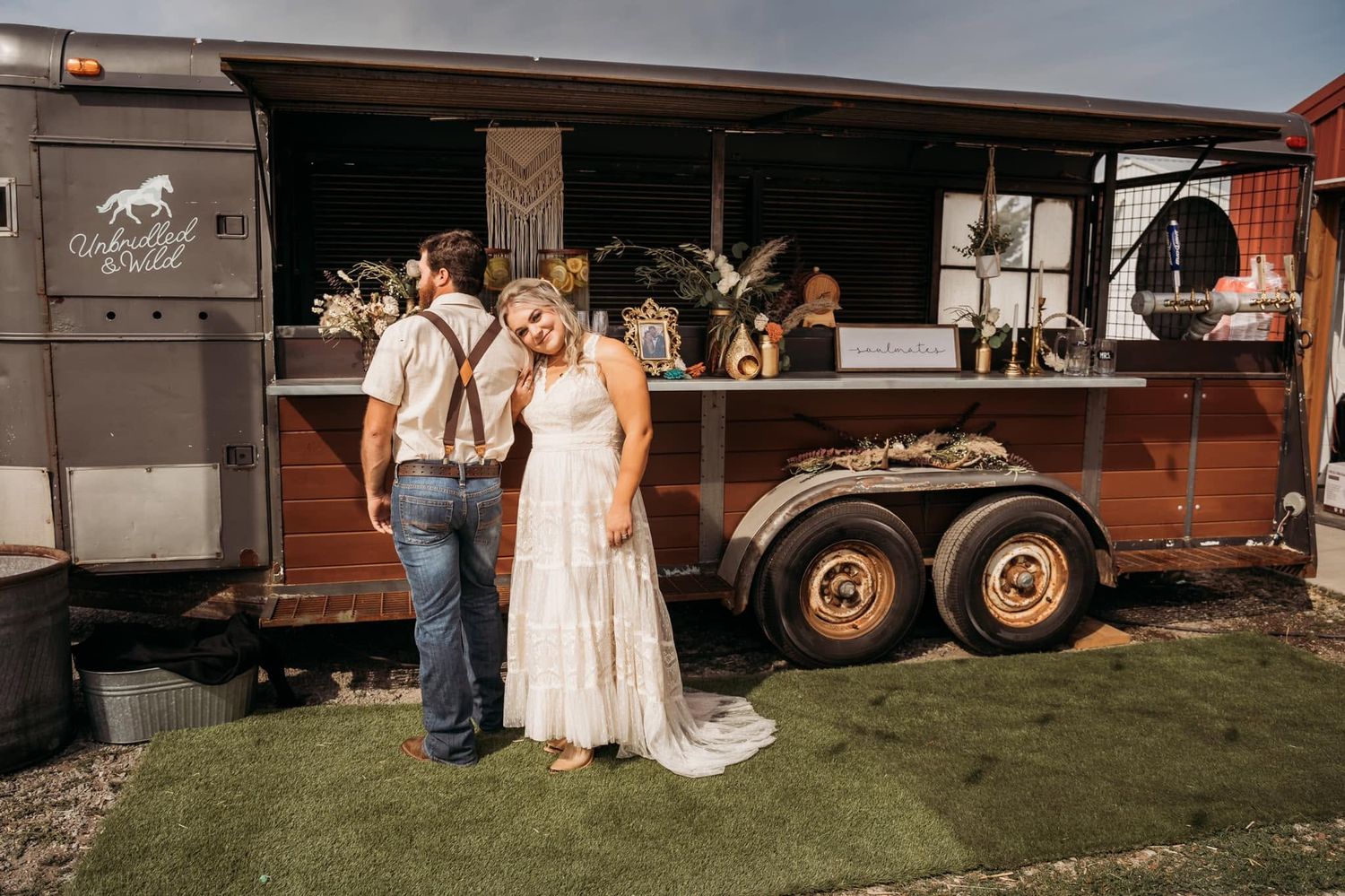 Bride and groom posing in front of rustic horse trailer bar at boho outdoor wedding.