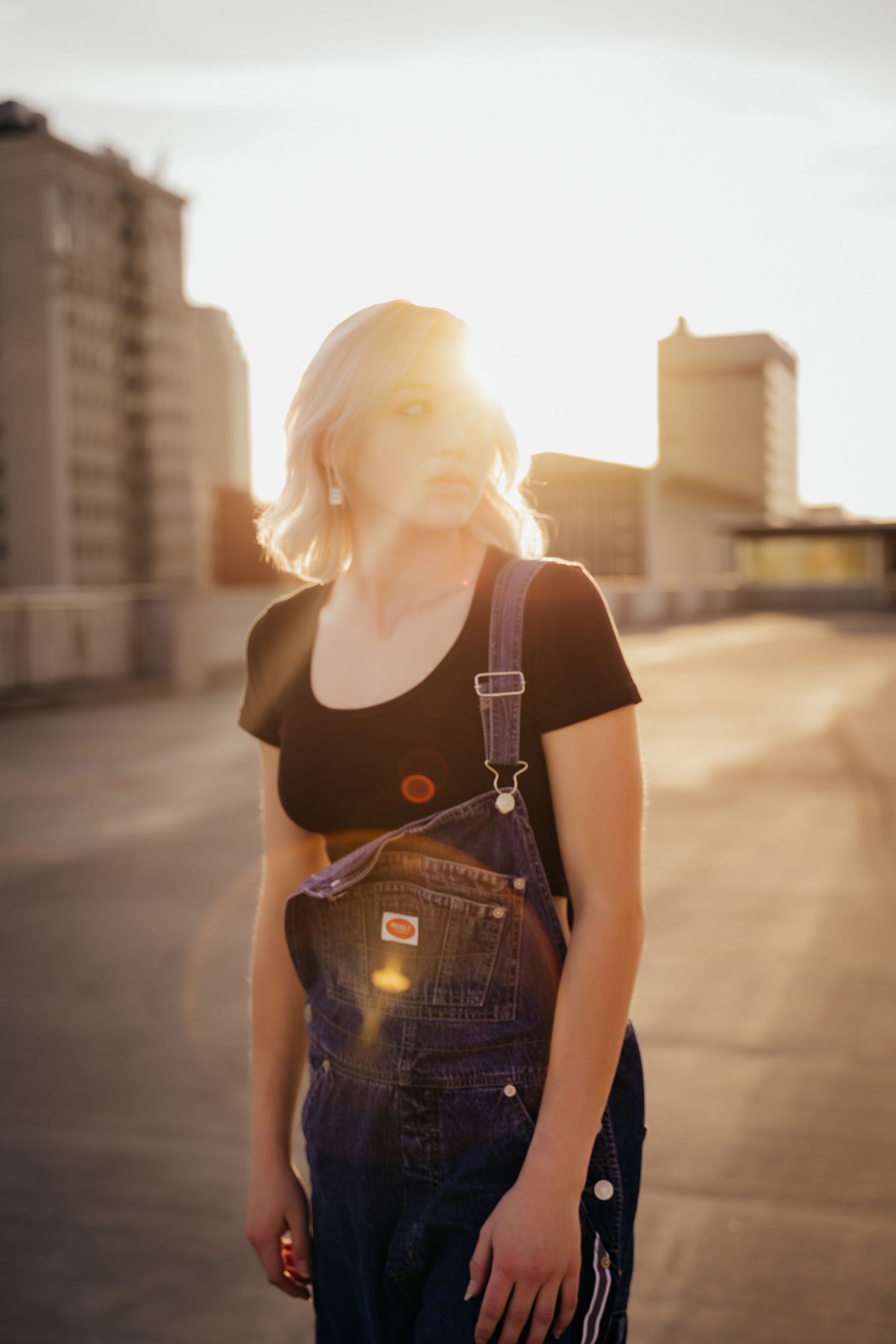 Blonde woman in denim overalls and black tee standing on rooftop at sunset with lens flare.