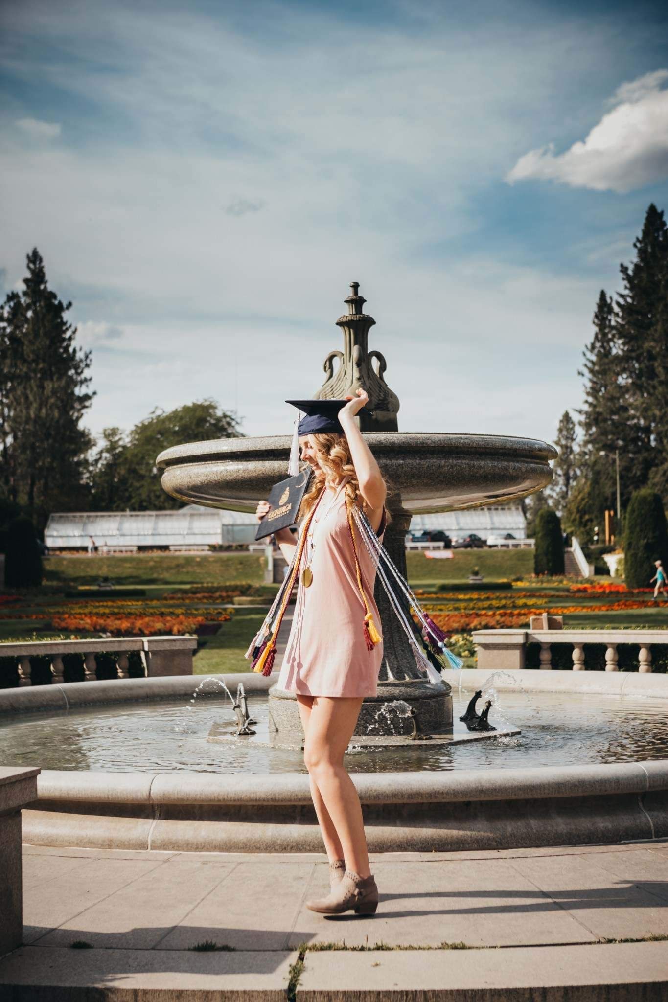 Blonde woman in pink dress celebrating graduation near an ornate fountain in a park.
