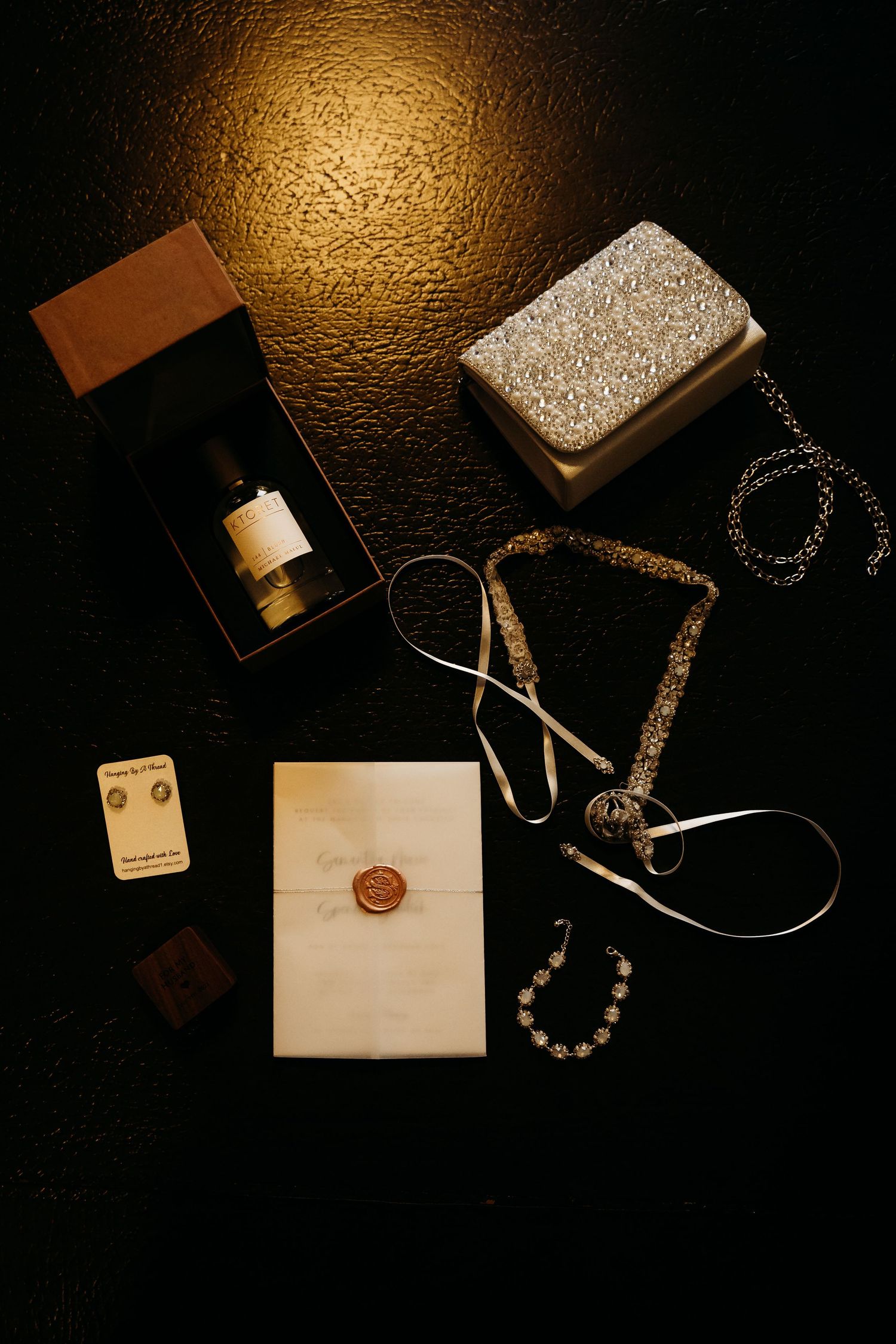 Flat lay of wedding accessories including perfume, wax sealed envelope, jewelry and sparkly clutch on dark surface.