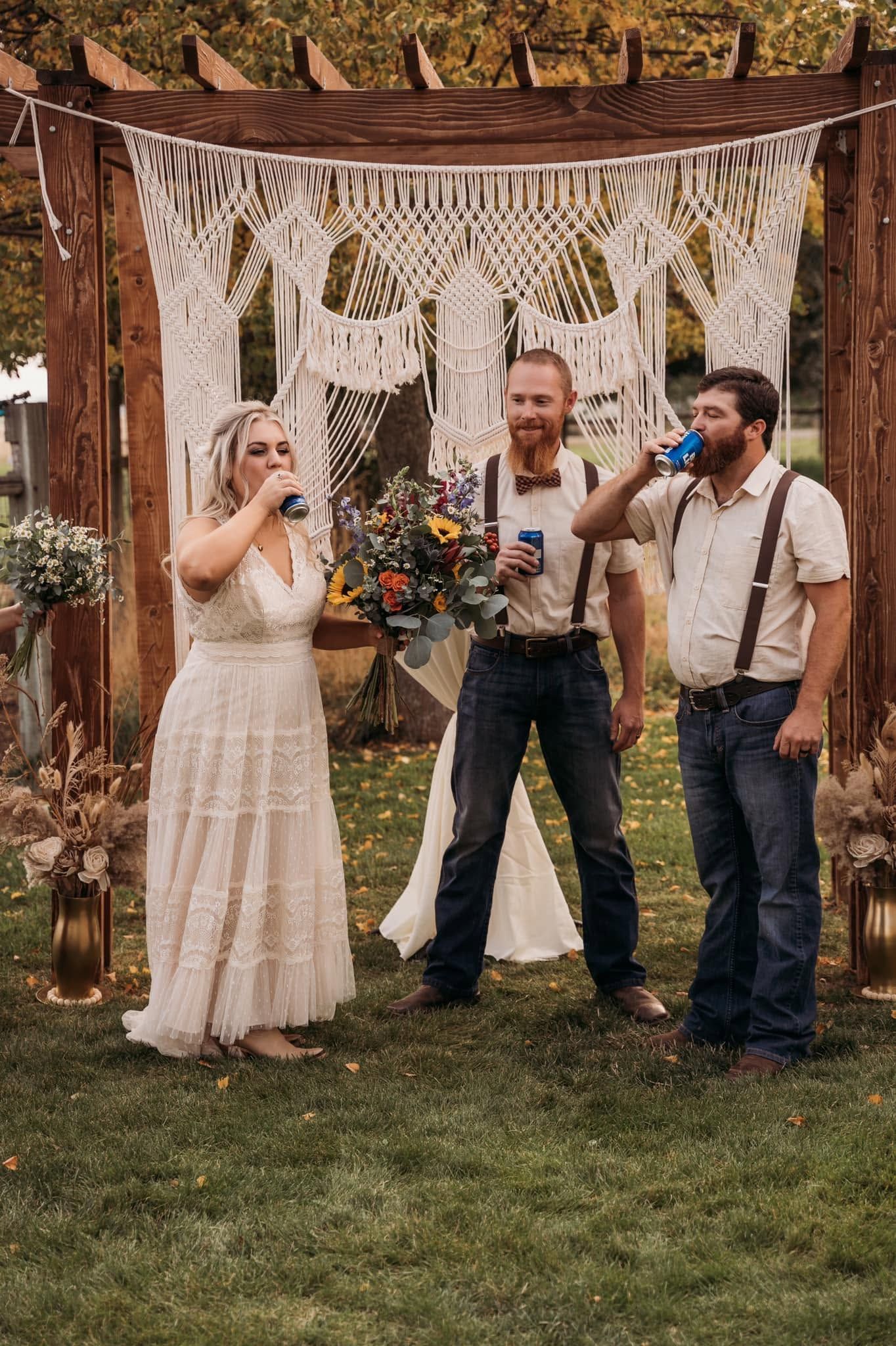 Bride, groom, and officiant toasting with cans under macrame arch at outdoor wedding ceremony.