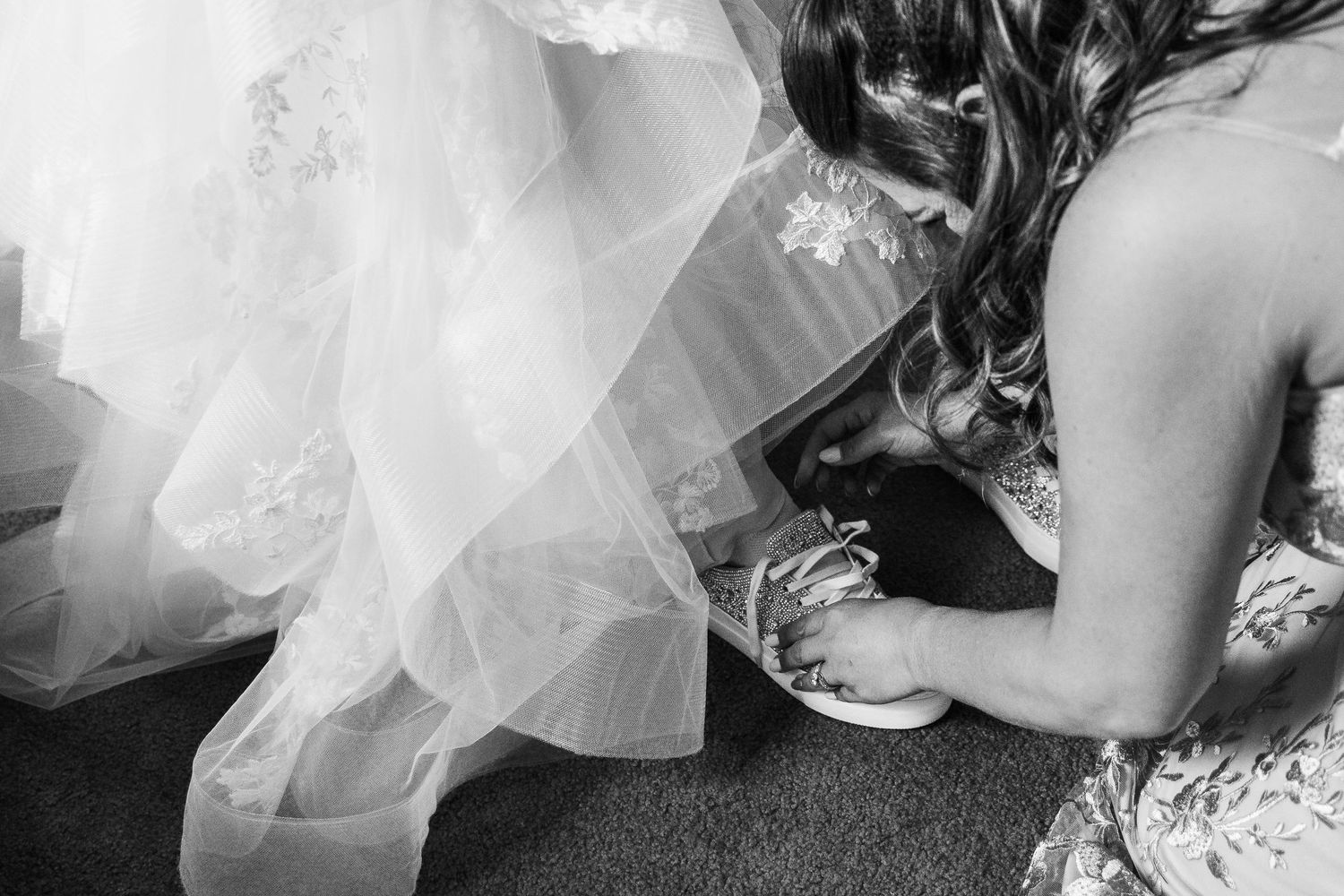 Black and white photo of bridesmaid helping bride put on crystal embellished sneakers.