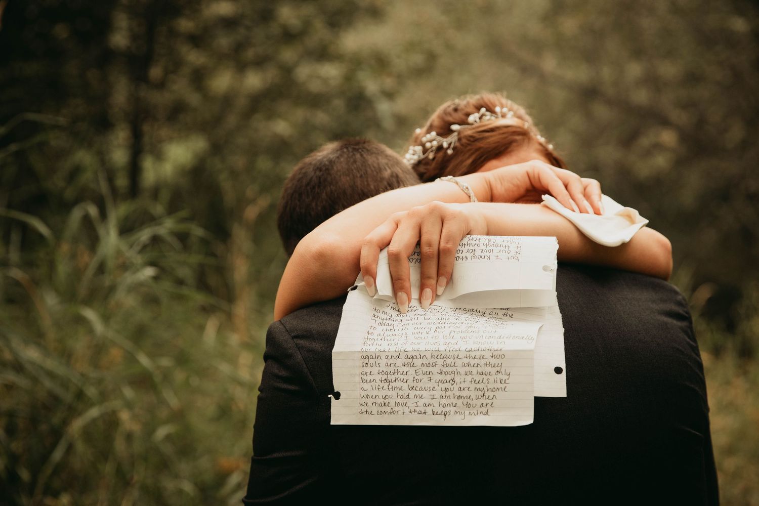 Bride hugging groom from behind holding handwritten wedding vows in a lush green forest.