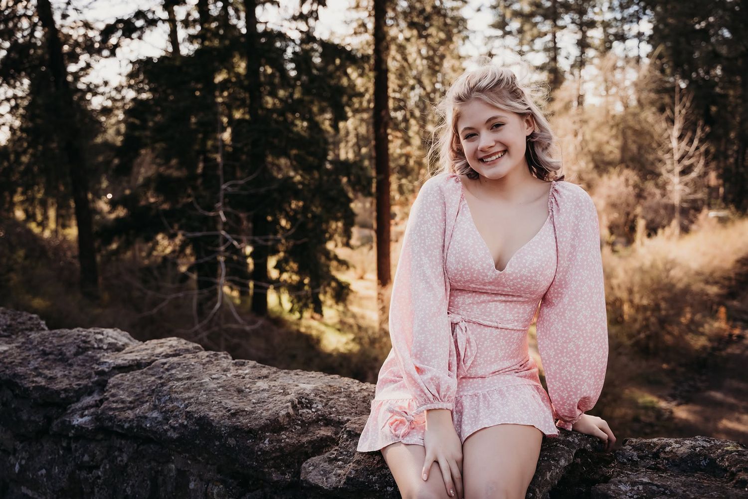 Smiling young woman in pink outfit sitting on rocks in sunlit forest.
