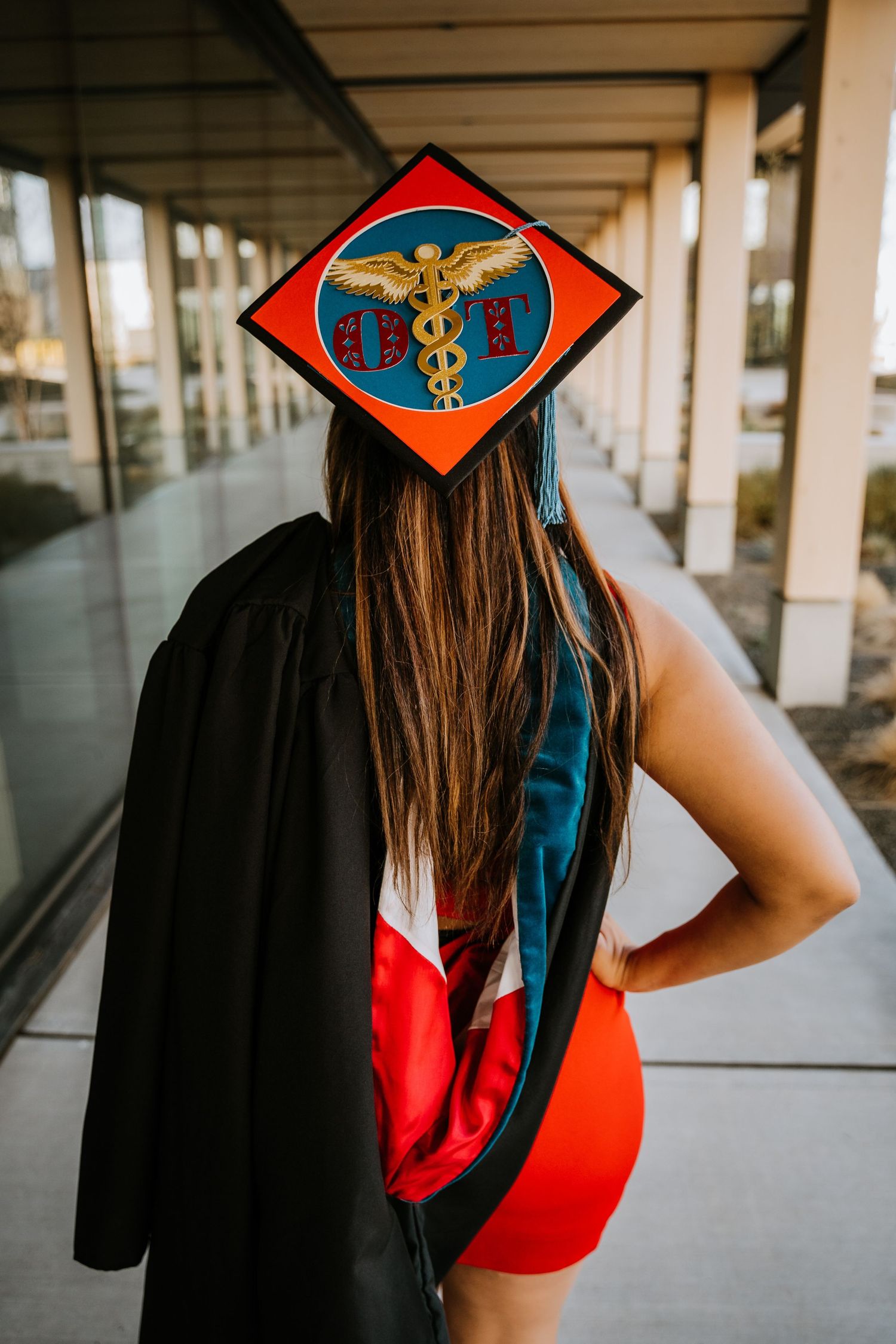 Graduate in red dress and black gown showing decorated medical graduation cap from behind.